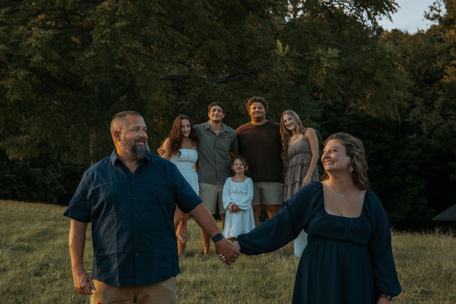 Smiling family of seven holding hands and walking together across a grassy hillside, with tall green trees in the background at sunset.