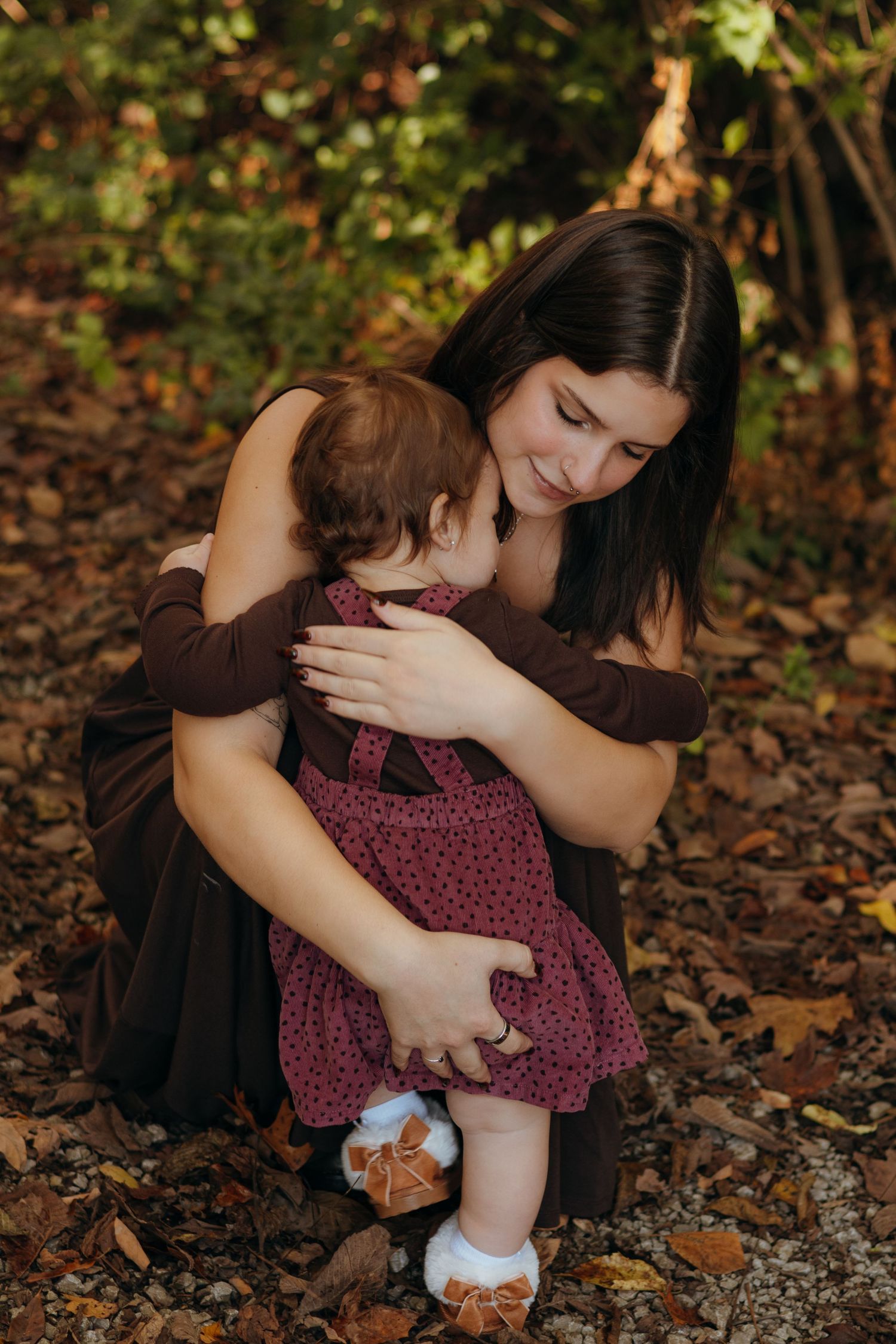 A smiling woman in a dark dress kneels on fallen autumn leaves, hugging a toddler in a burgundy pinafore outdoors.