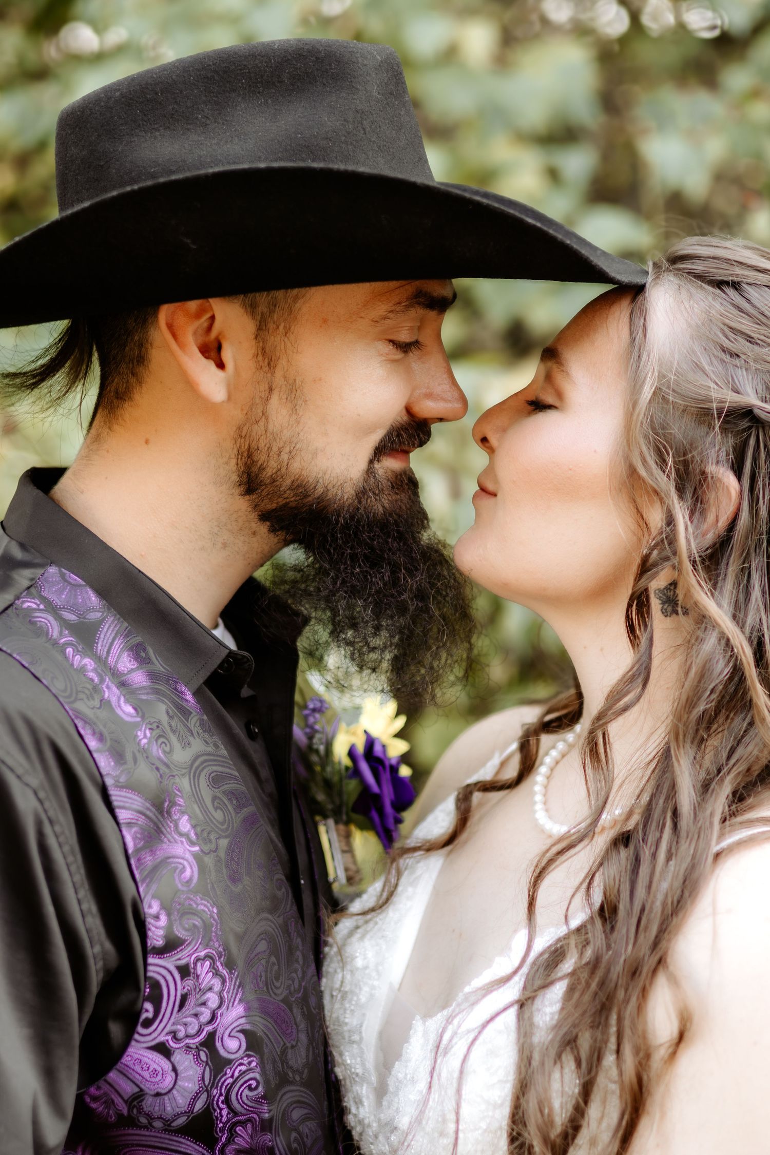 Bearded groom in a black cowboy hat and purple paisley vest nose to nose with his bride in a white lace gown and pearl necklace.
