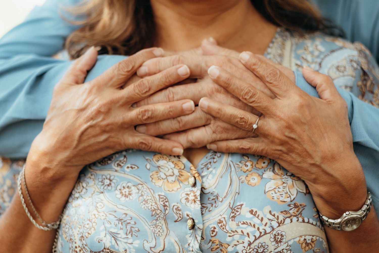 Close-up of an elderly woman's wrinkled hands clasped over a younger person's hands, wearing a floral blue top and silver bracelet.