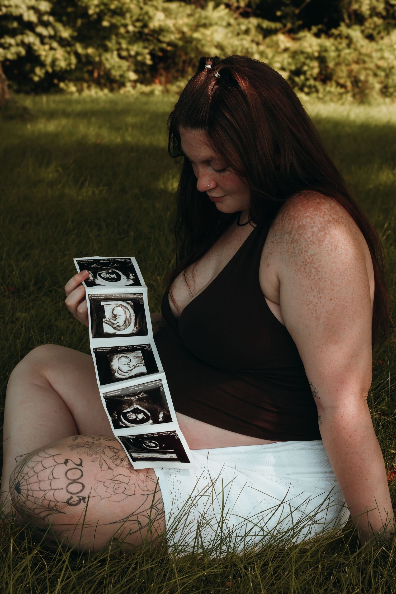 Woman with long dark hair sits on green grass outdoors, gazing down at a strip of black-and-white ultrasound photos in her hand.