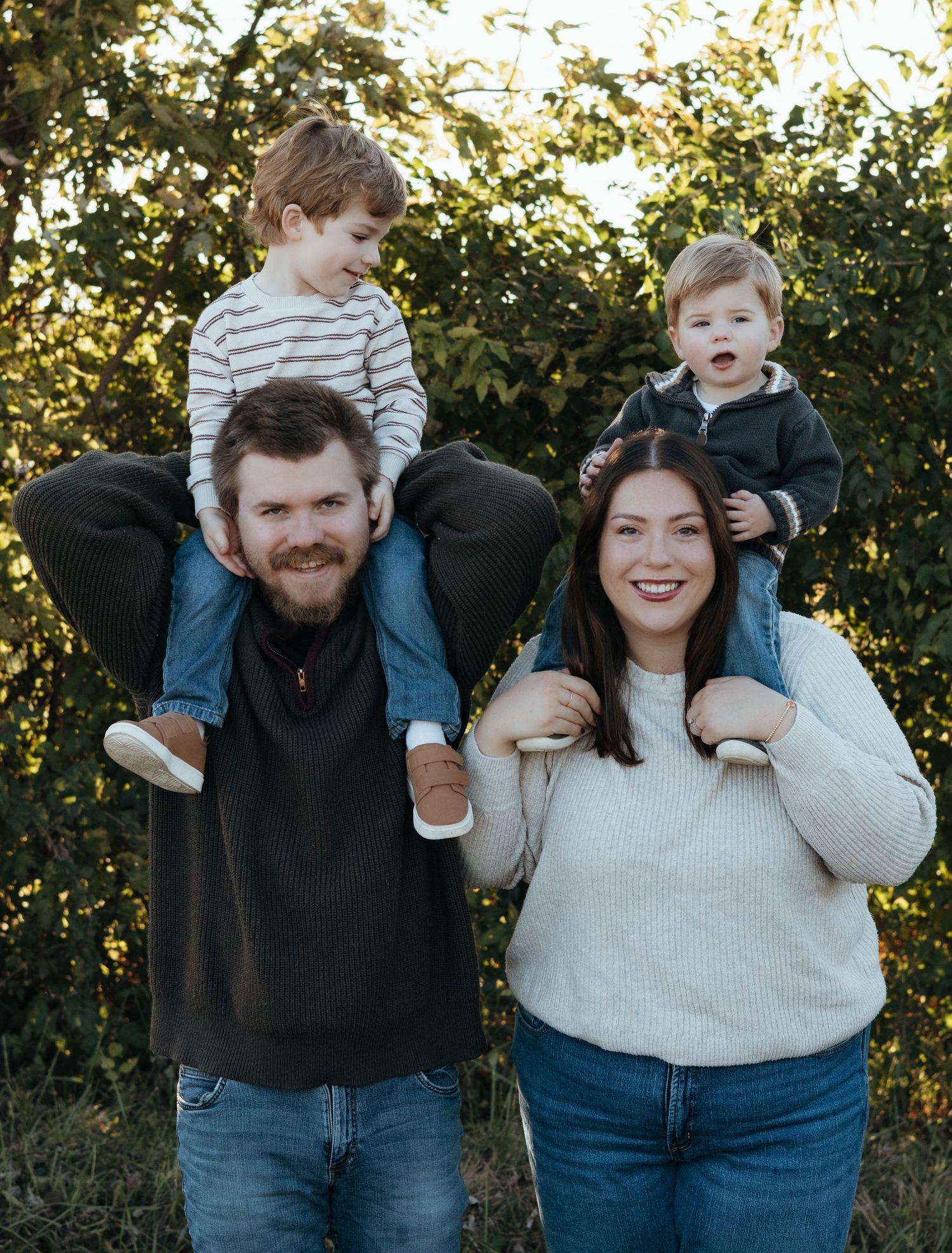 Smiling family portrait outdoors with a bearded man and a woman each carrying a young child on their shoulders amid green foliage.