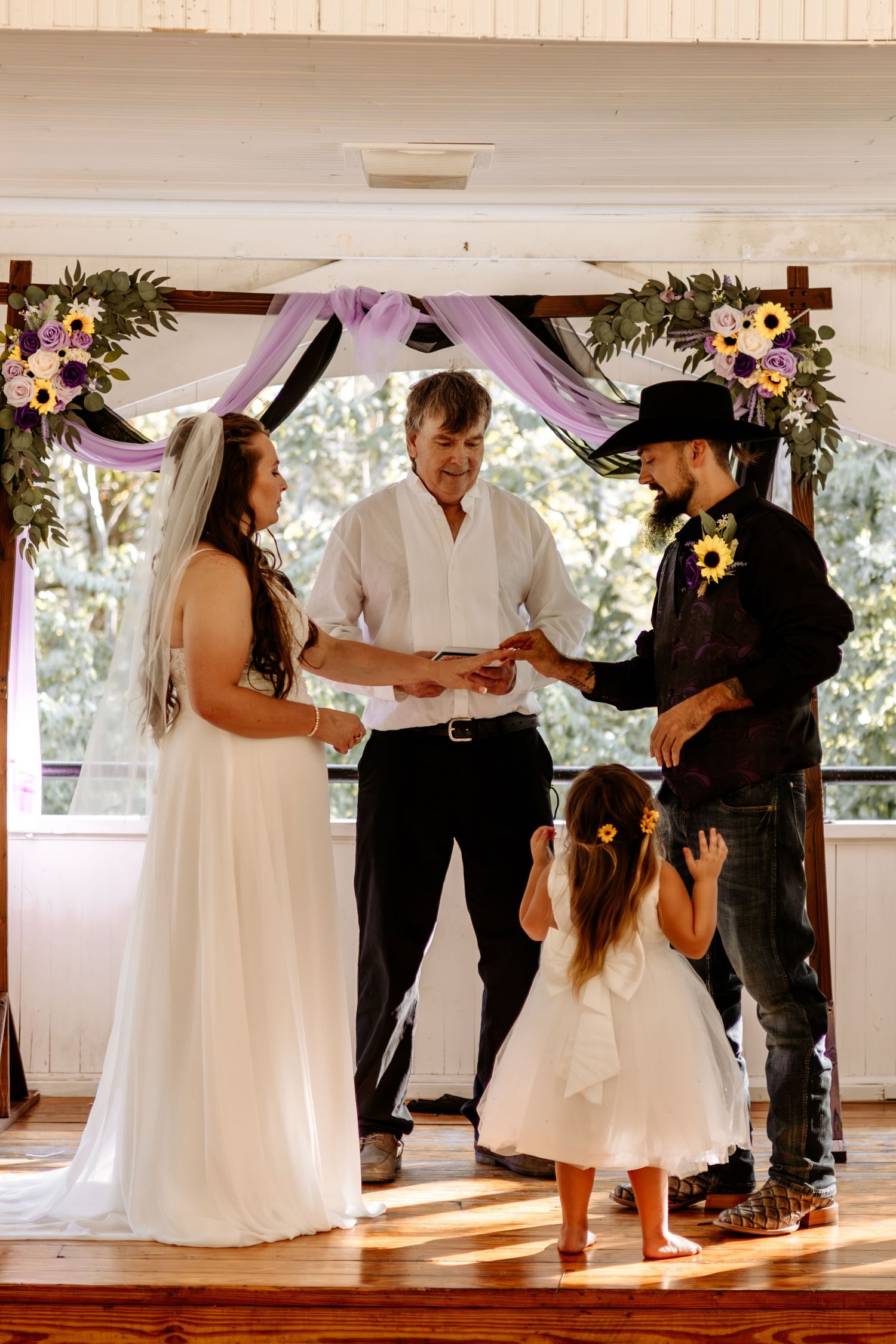 Bride in a white gown and groom in a black hat exchange vows under a floral arch while an officiant and flower girl look on.
