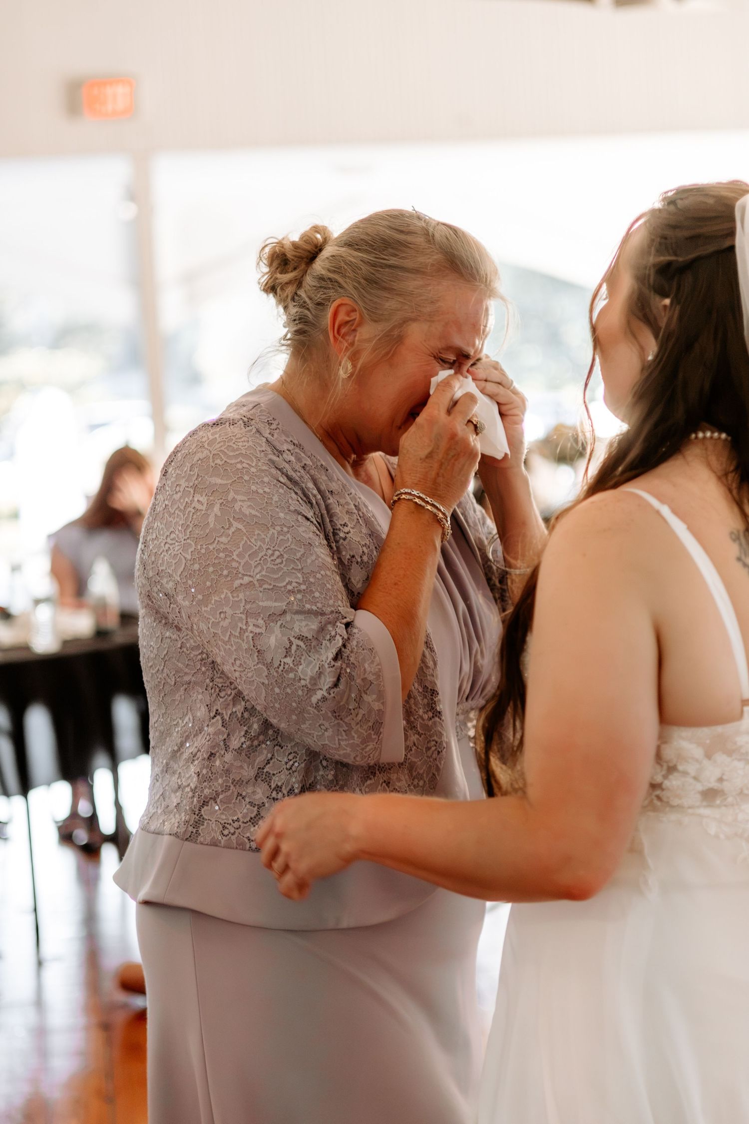 Emotional mother in a lace gray dress wipes tears with a tissue while embracing a bride in a white wedding gown at the reception.