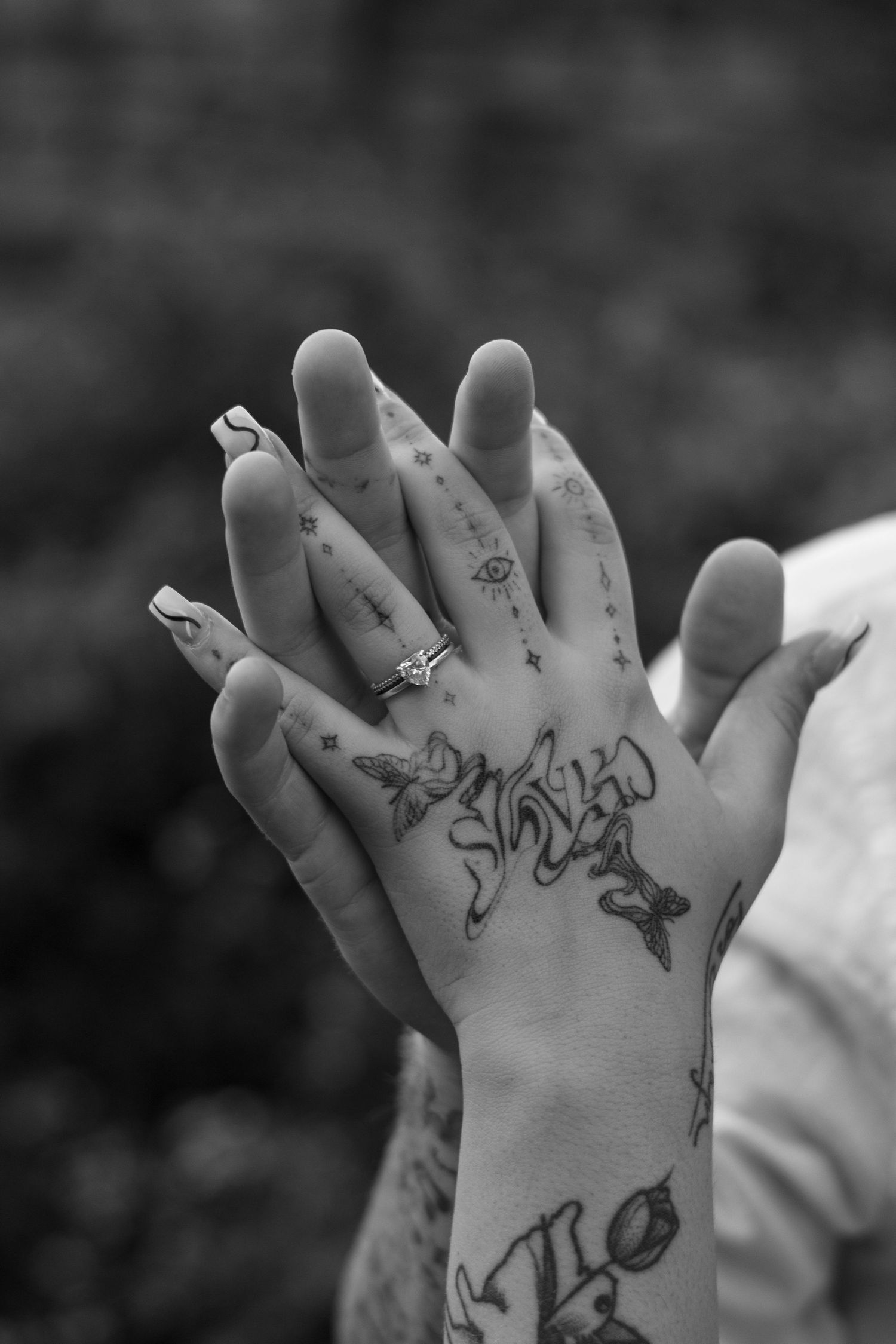 Black and white photo of two clasped hands, one with intricate tattoos and a diamond engagement ring, against a blurred outdoor background.