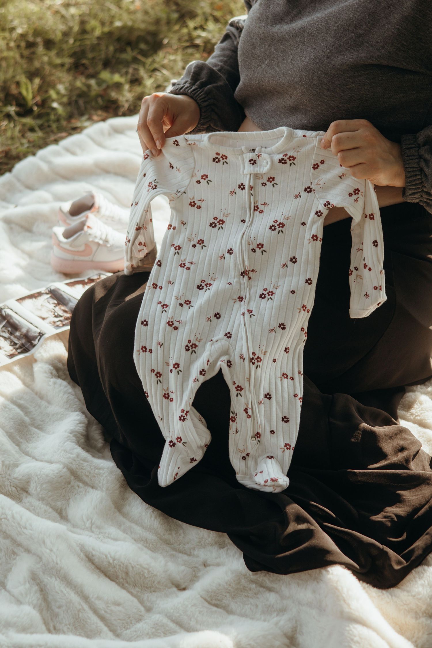 Person in dark skirt sitting on a cozy blanket holds up a white footed baby sleeper with a red floral print.