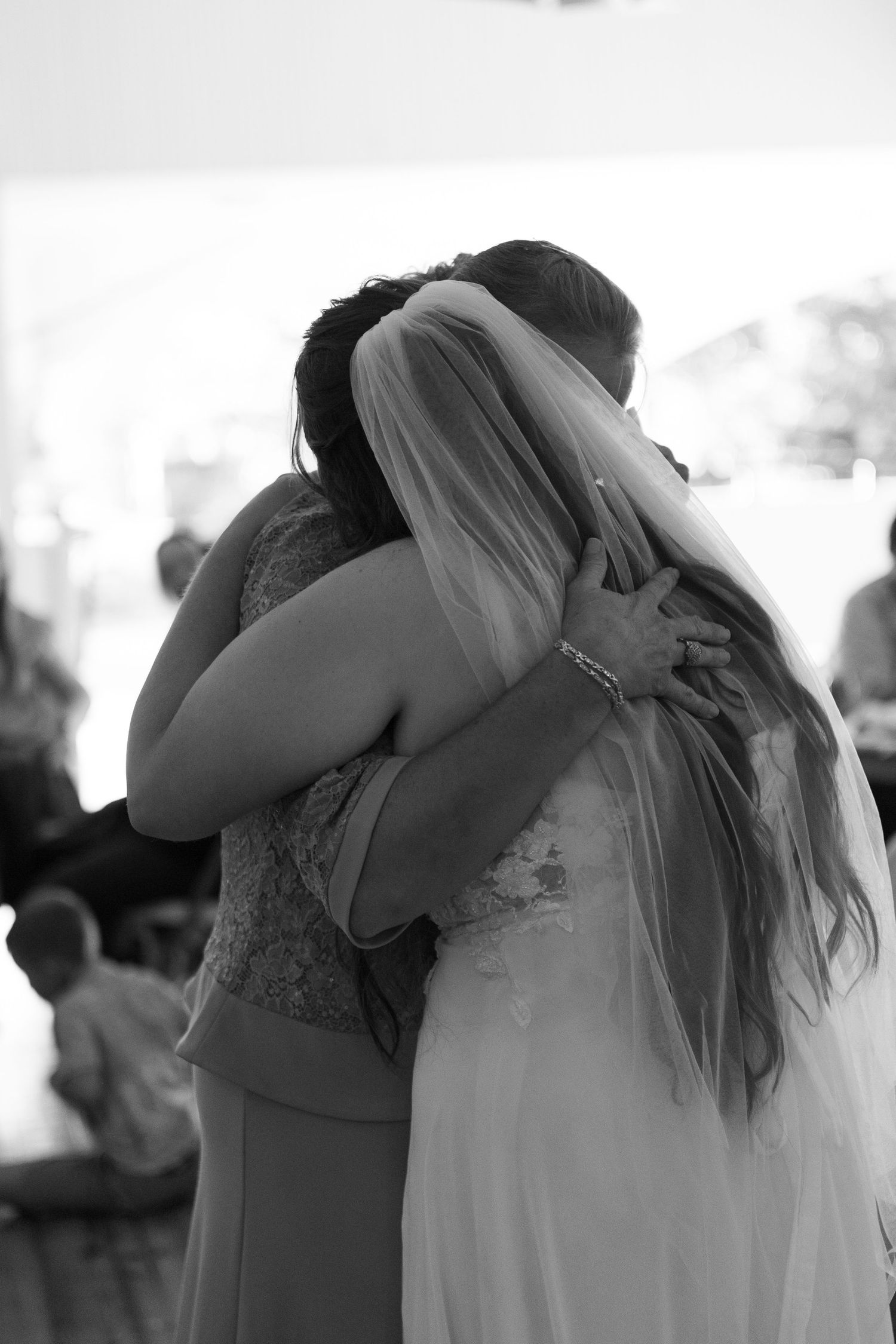 Black and white photo of a bride in a veil embracing a woman at a wedding reception in a warm, emotional hug.