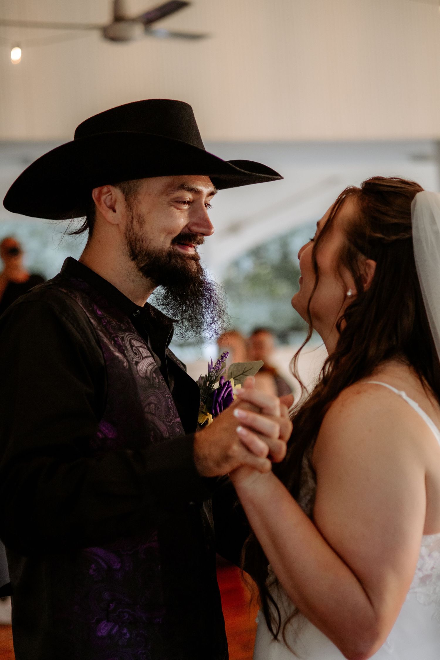 Smiling groom in a black cowboy hat and purple vest holds hands with his veiled bride in a white dress during a joyful wedding dance.
