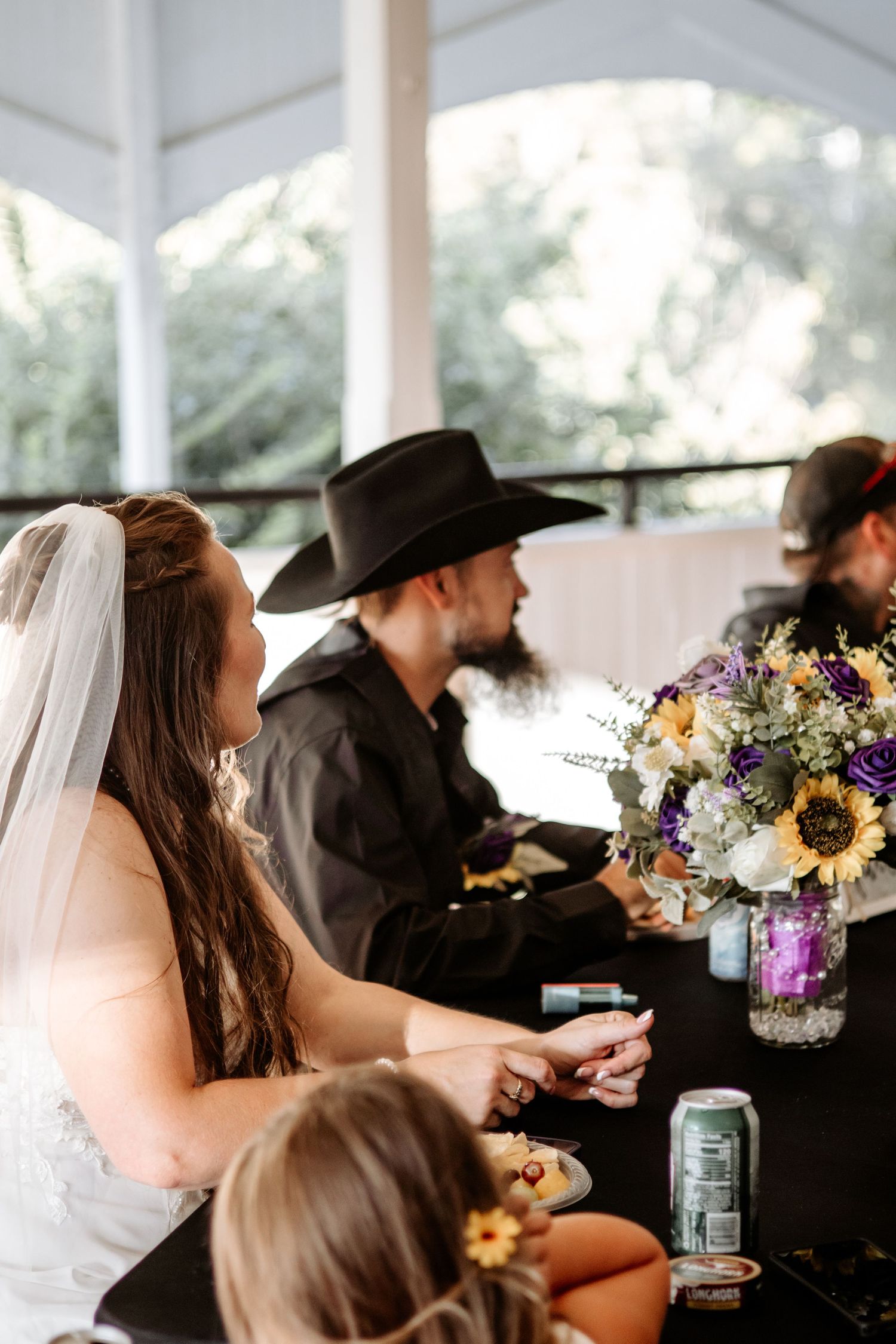 Bride in a veil and white gown sits at a reception table with a man in a black cowboy hat beside a sunflower and purple floral centerpiece.