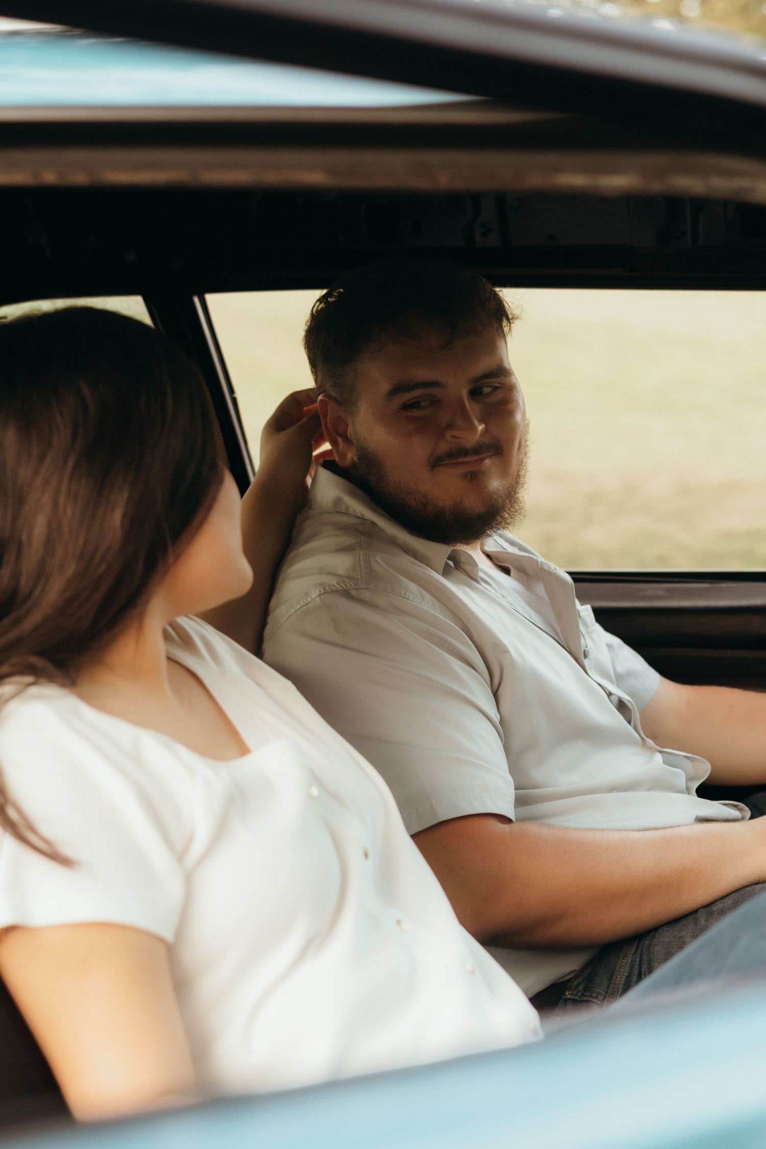Smiling bearded man in a button-up shirt sits in a vintage car while a woman in a white top touches her hair beside him.