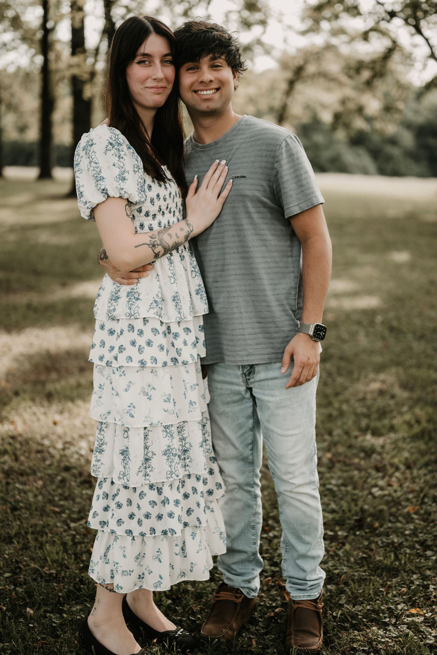Smiling couple poses outdoors under trees, she in a ruffled floral dress and he in a gray t-shirt and jeans.