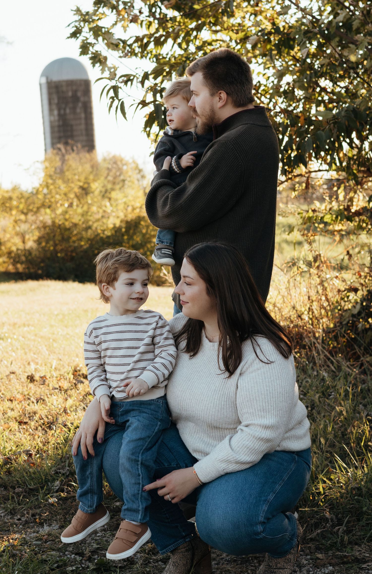 Family of four posing outdoors in autumn light, a woman kneeling with a young boy while a man holds a toddler, with a silo behind.