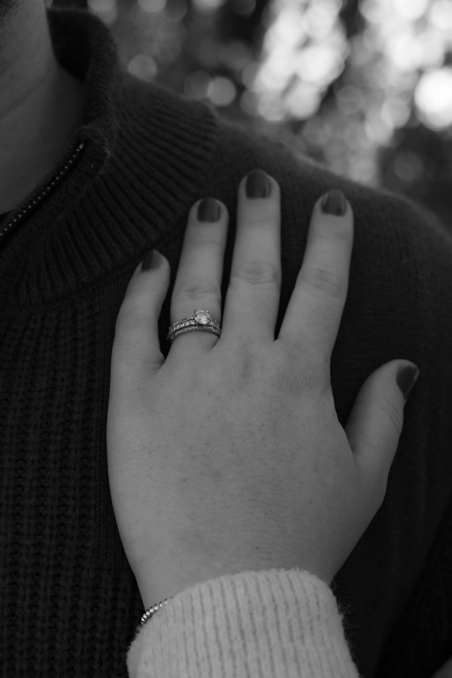 Black and white close-up of a hand with dark nail polish and a diamond engagement ring resting on a person's knit sweater shoulder.