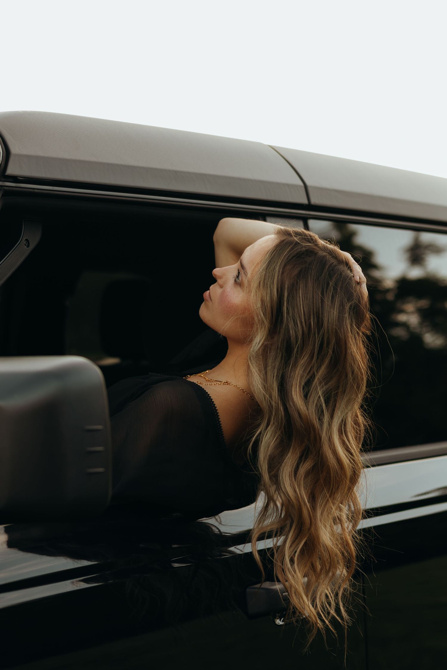 Woman with long wavy hair leans out of a black vehicle window, resting her arm on her head and gazing upward at dusk.