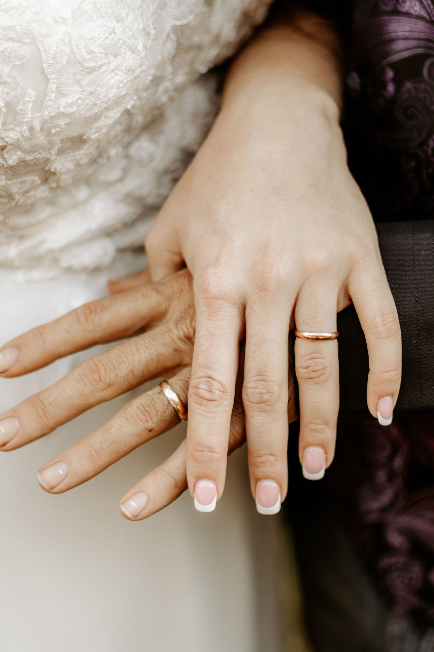 Two hands with wedding rings and French manicures rest together, with a bride in a white lace gown and a person in a purple jacket.