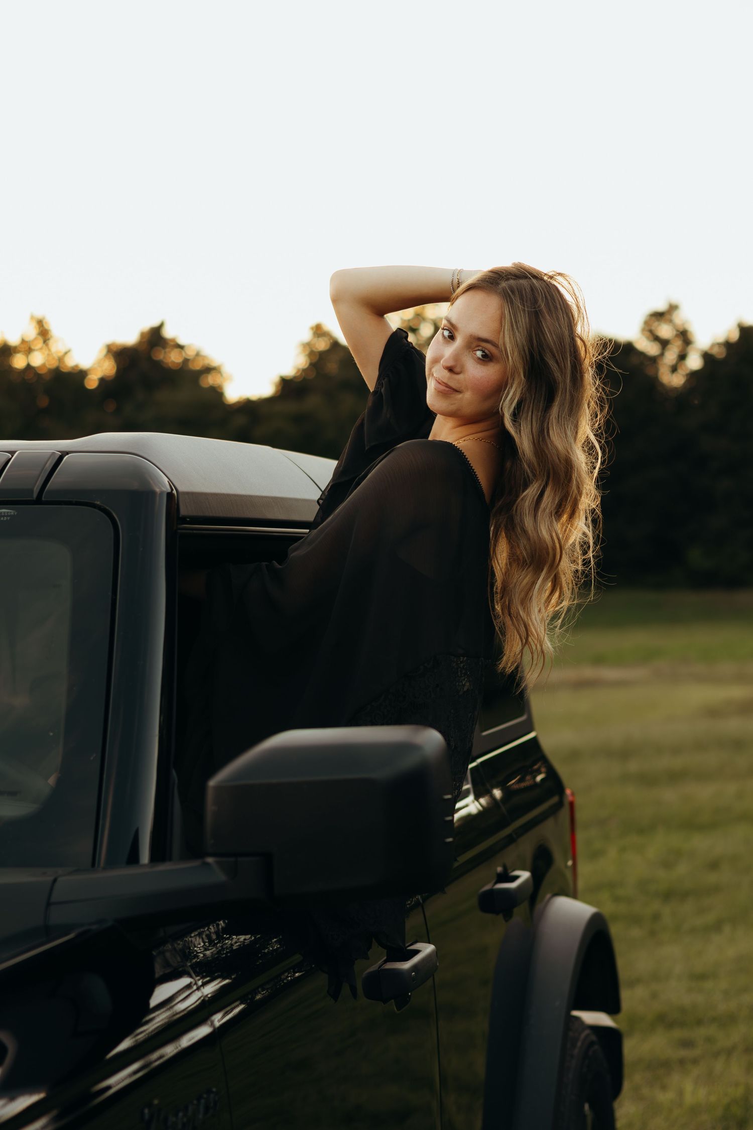 Woman with long wavy hair leaning out of a black Jeep window at golden hour, with a grassy field and trees behind her.