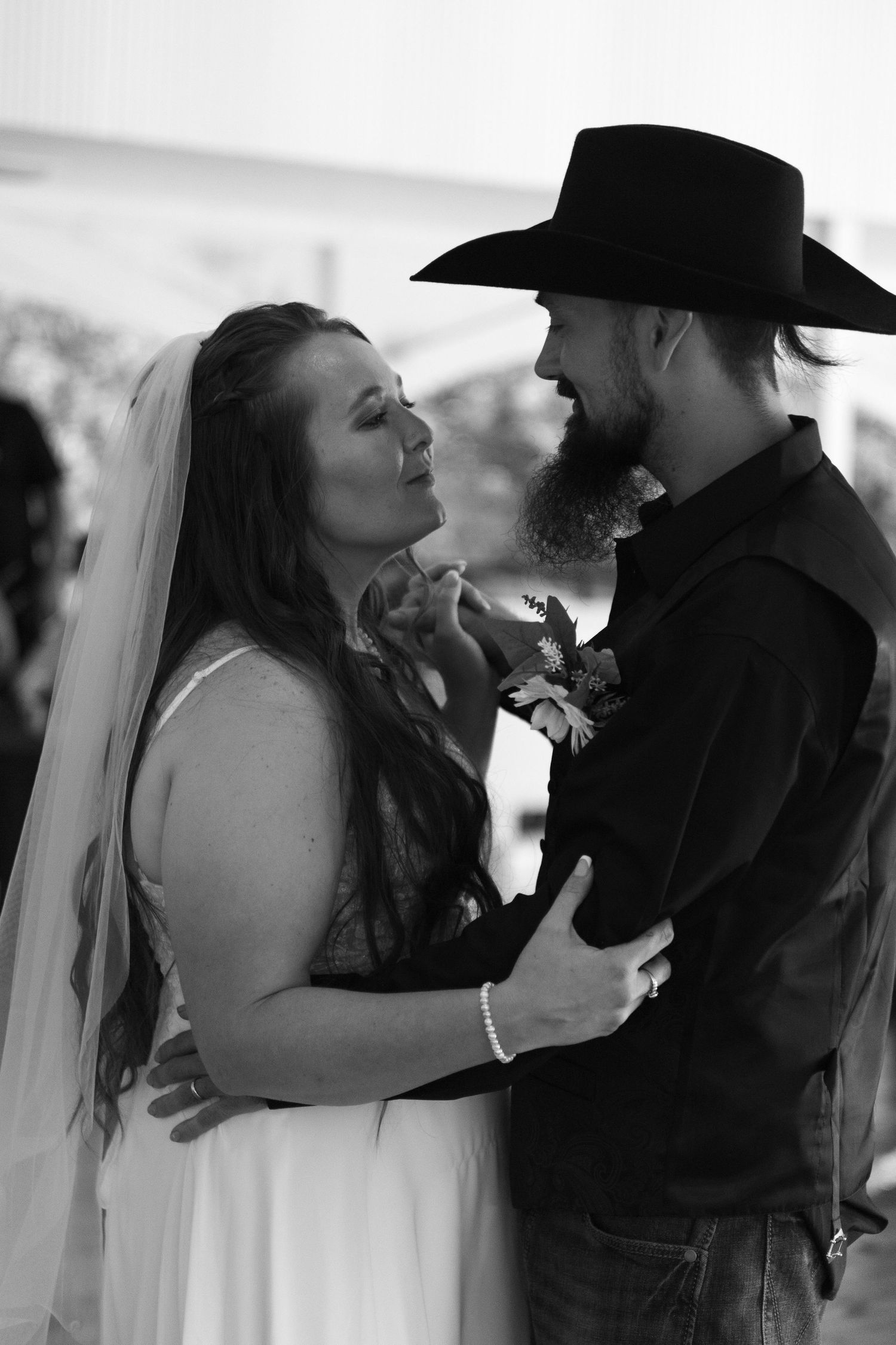 Black and white photo of a bride in a veil and a bearded groom in a cowboy hat sharing a close first dance at their wedding.