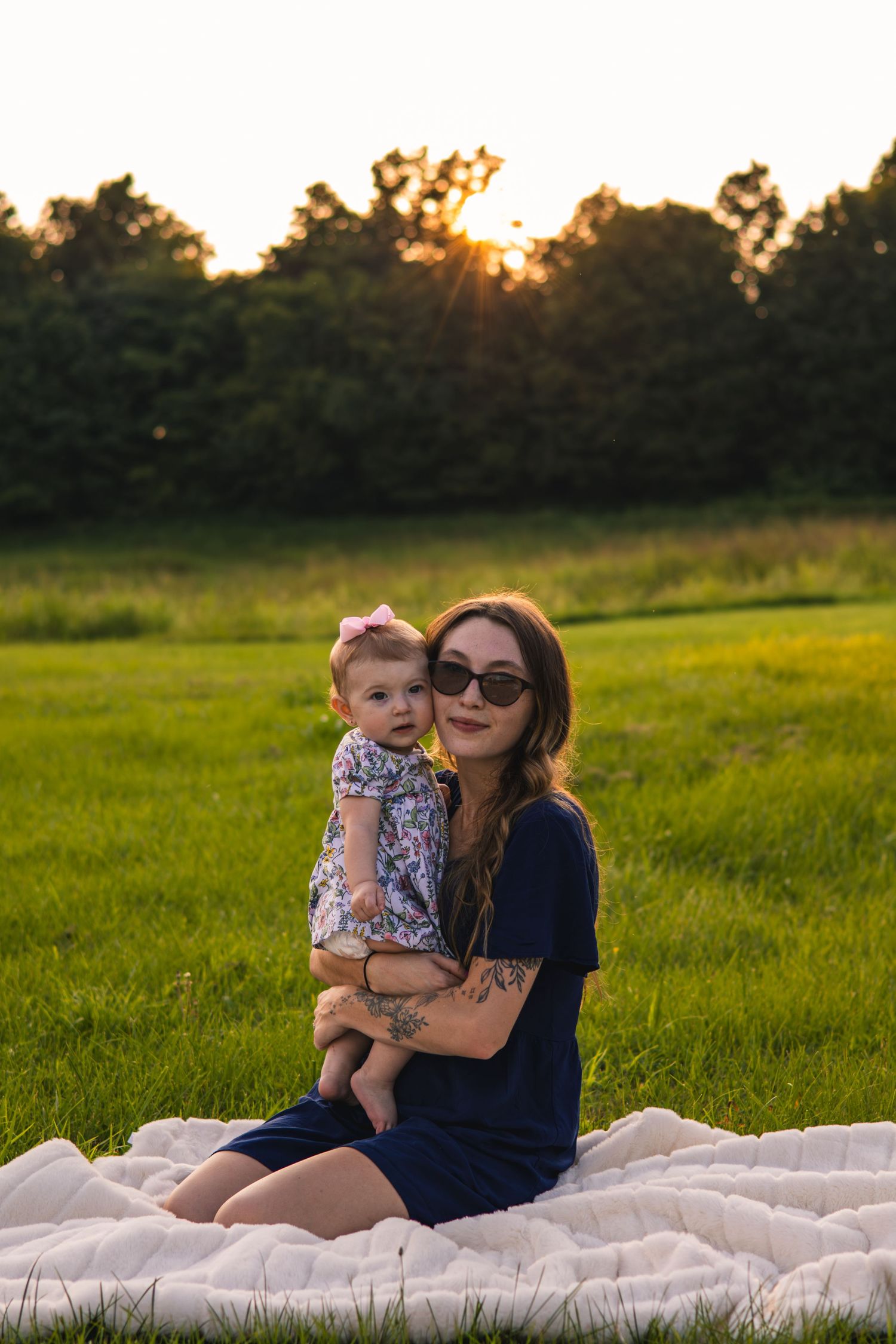 Woman in sunglasses and a navy dress holds a baby in a floral outfit while sitting on a white blanket in a grassy field at sunset.