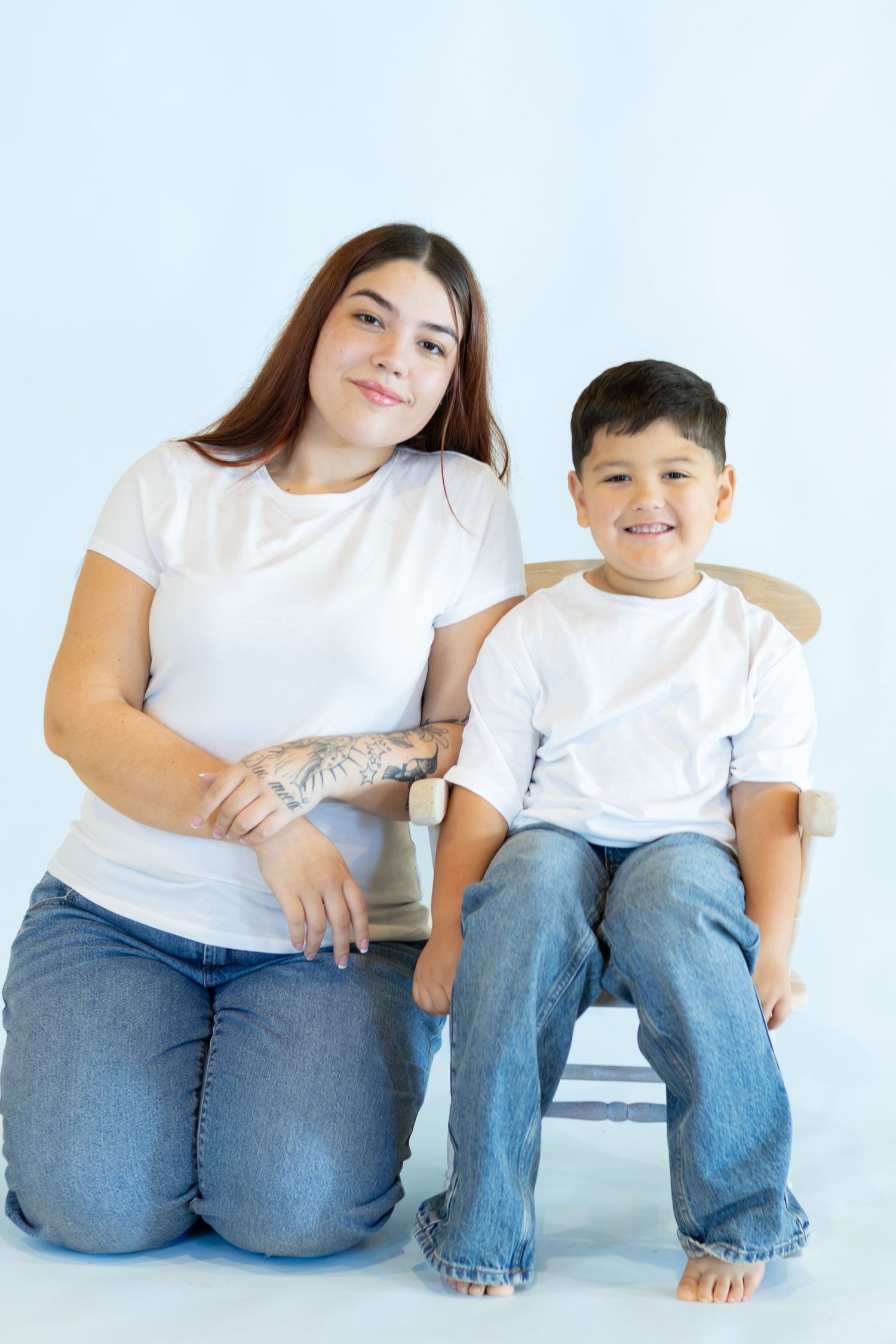 A smiling young woman with a forearm tattoo kneels beside a smiling boy seated on a chair, both in white tees and jeans.