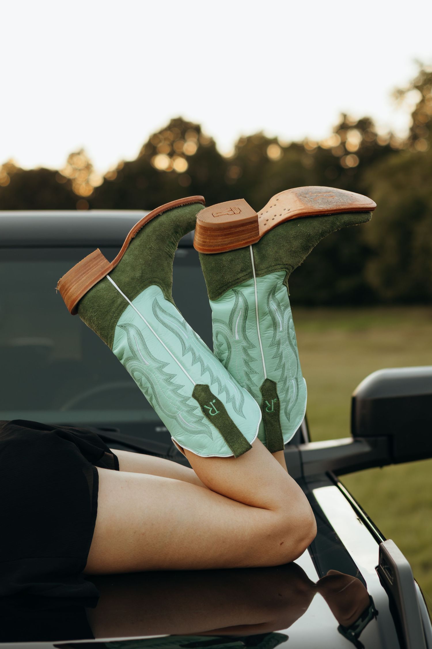 Woman wearing green and mint cowboy boots with stitched details, legs propped up on a truck hood at sunset in a grassy field.