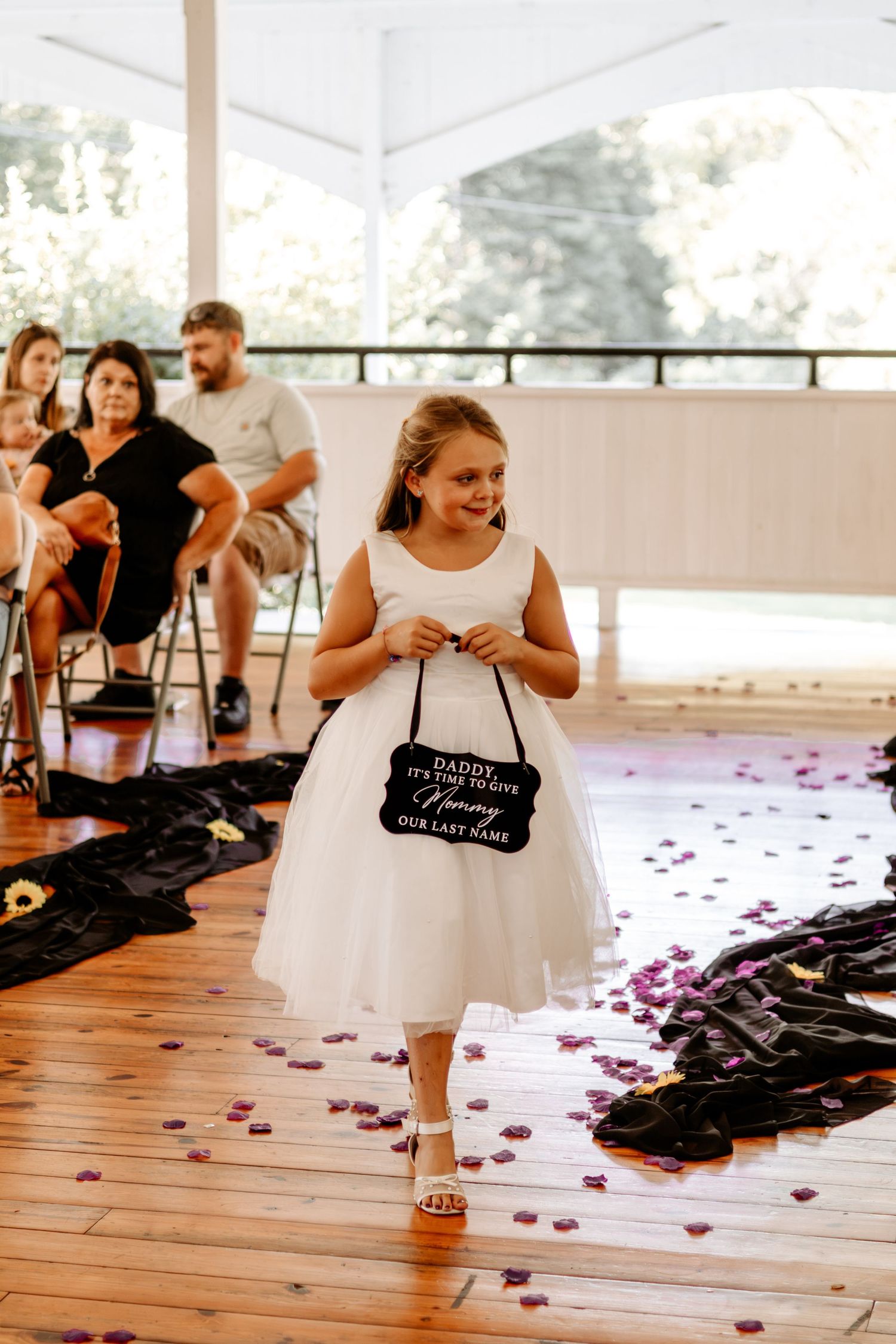 Smiling flower girl in a white dress holds a sign reading Daddy, it's time to say hello Mommy, as she walks down a petal-covered aisle.