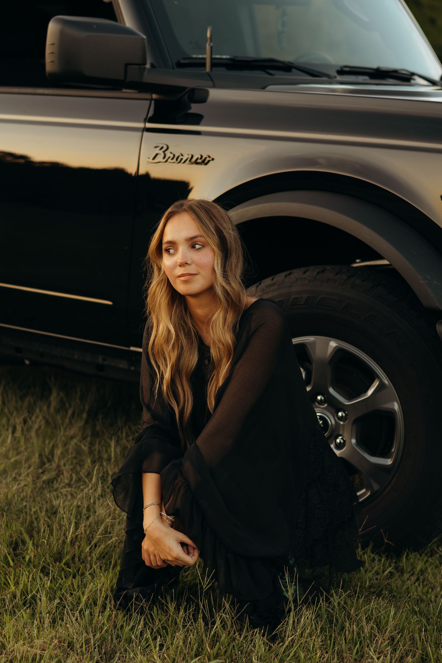 Woman with long wavy hair in a black dress crouches in the grass beside a black Ford Bronco SUV at golden hour.