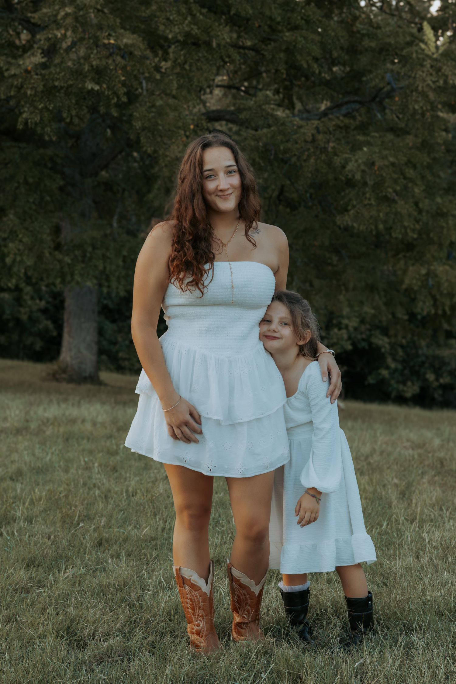 Woman in a white strapless dress and brown cowboy boots stands beside a smiling girl in a white dress on a grassy lawn under trees.