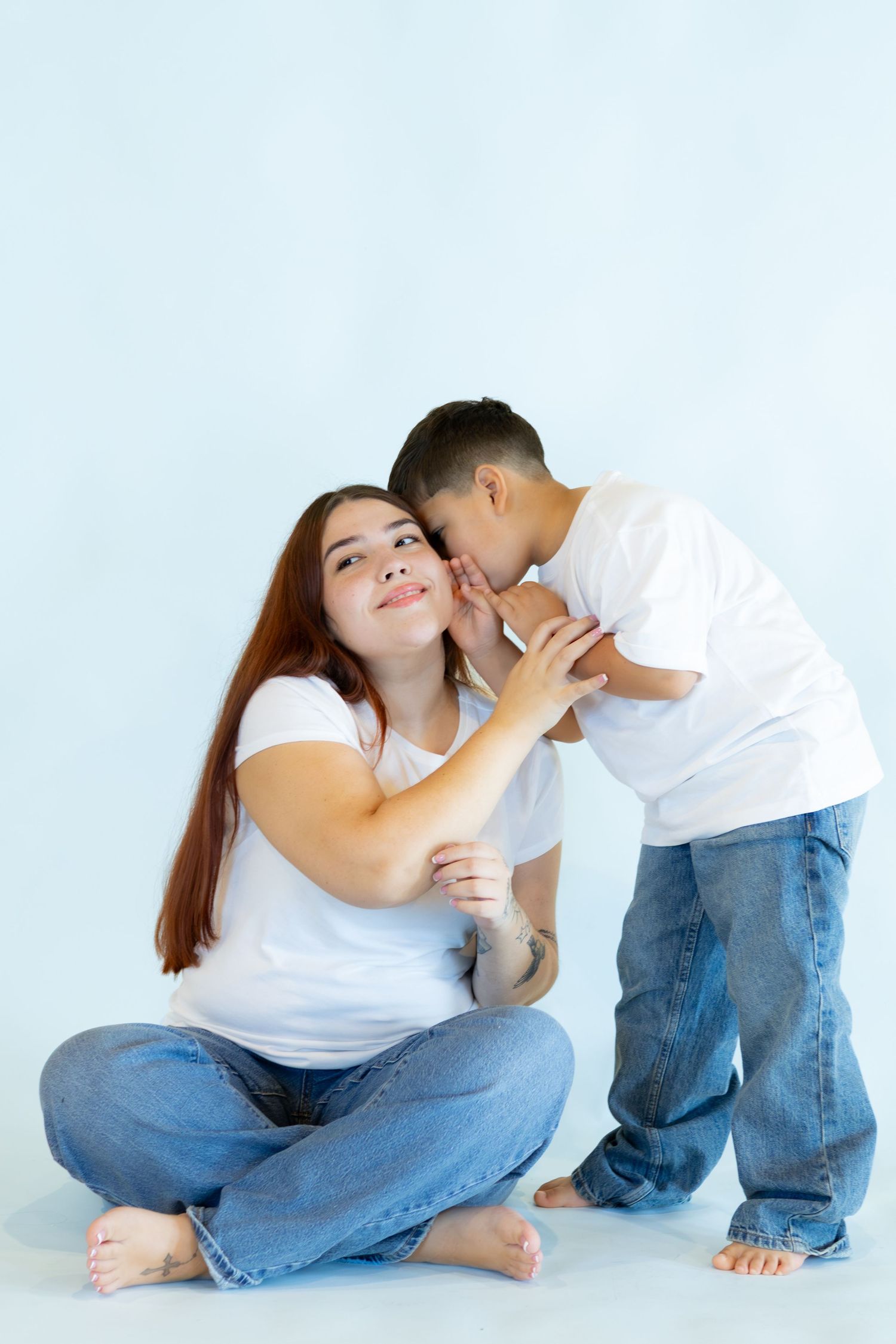 A smiling woman with long auburn hair sits barefoot on the floor in jeans and a white tee while a young boy whispers in her ear.