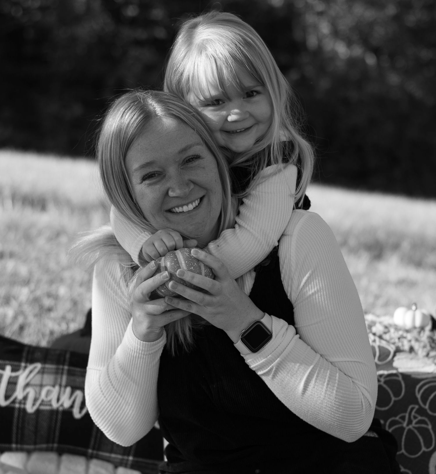 Black and white portrait of a smiling woman being hugged from behind by a little girl during an outdoor picnic.