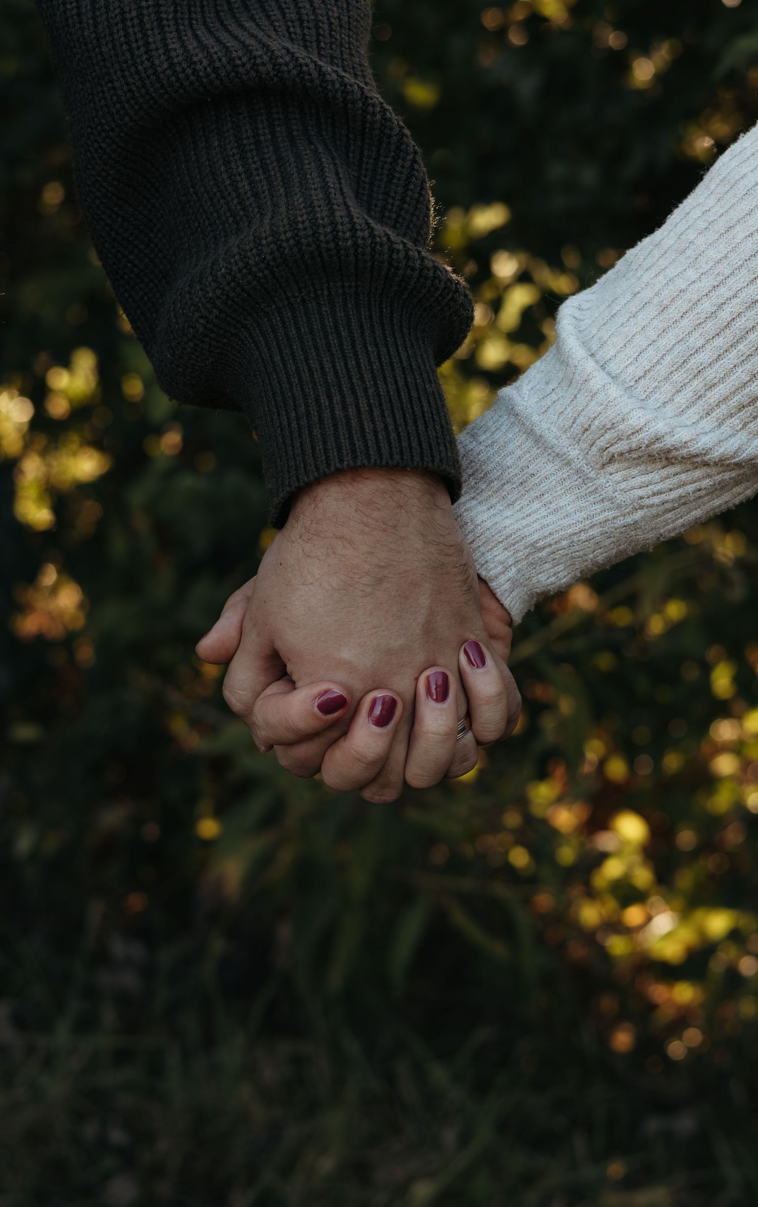 Two people hold hands outdoors in cozy knit sweaters, one with burgundy nail polish, against a softly blurred autumn foliage background.
