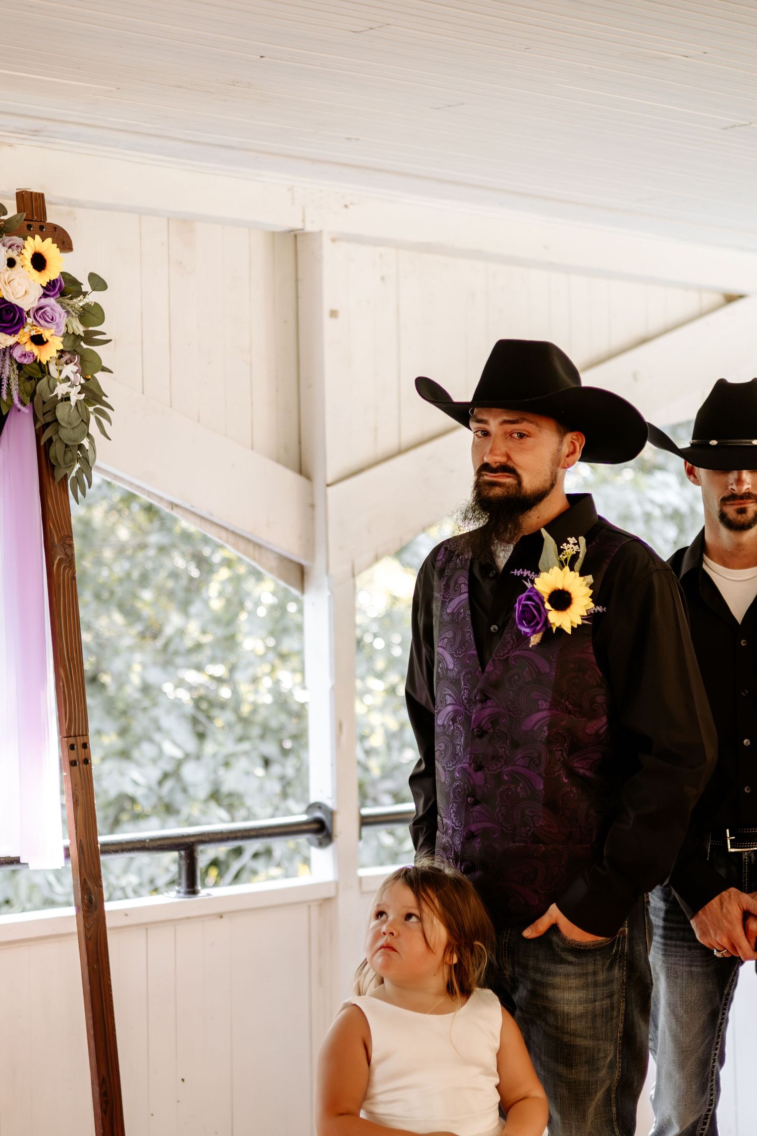 Groom in a black cowboy hat and purple vest with a sunflower boutonniere stands at a rustic wedding as a young girl looks on.
