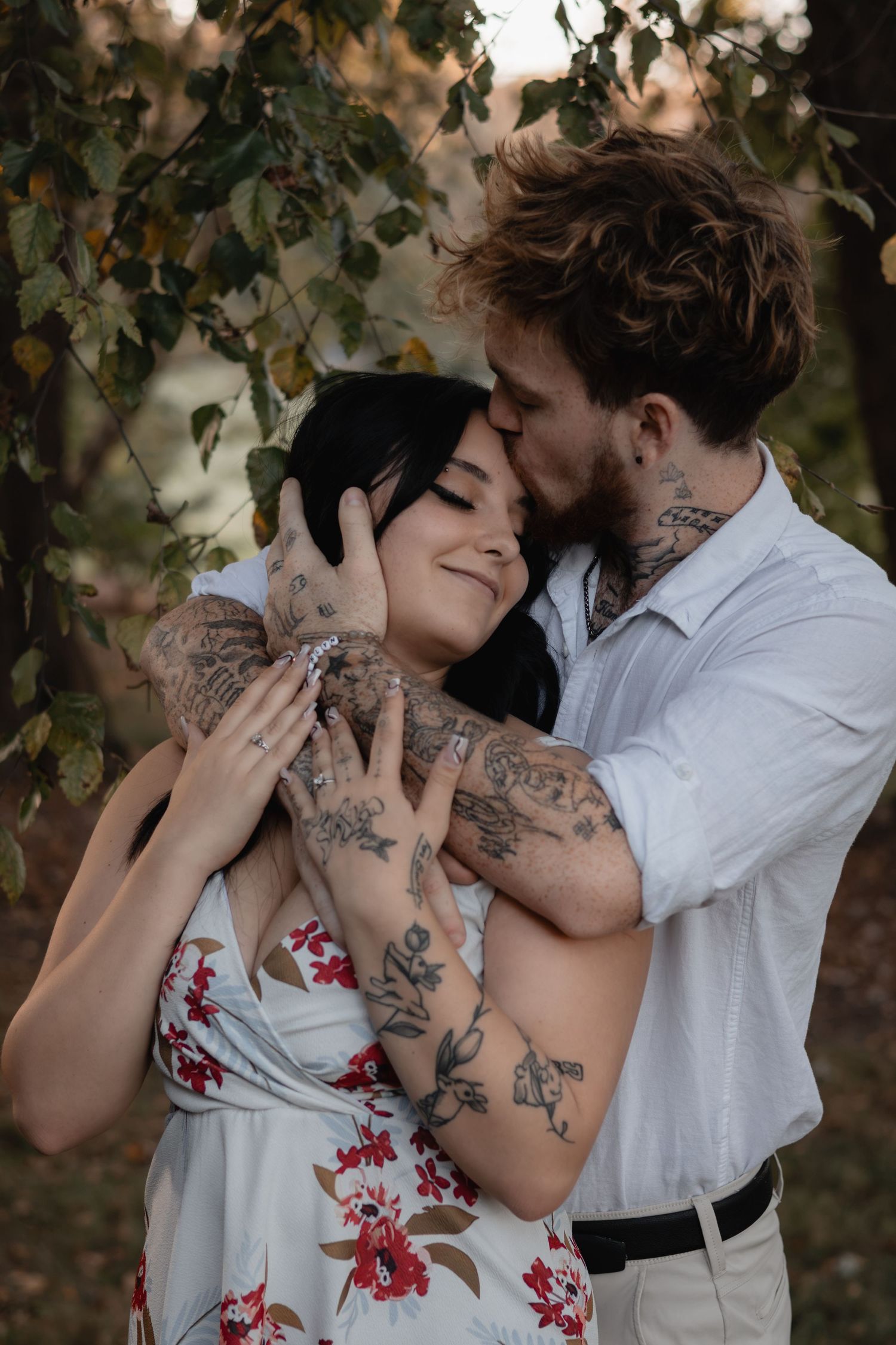 Tattooed couple embracing outdoors under trees as a bearded man kisses the forehead of a smiling woman in a floral sundress.