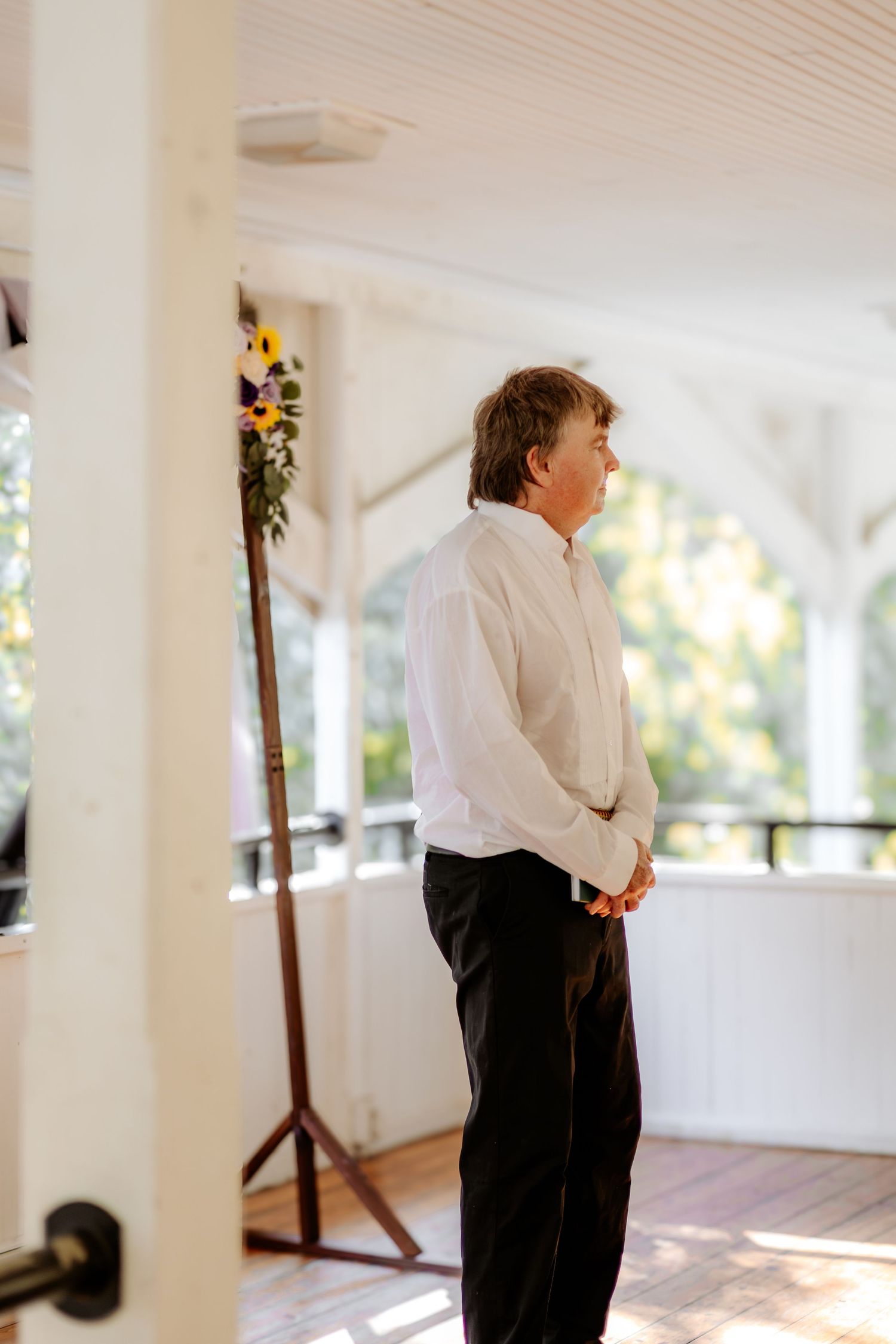 Man in a white shirt and black pants stands with hands clasped on a sunlit wooden pavilion porch beside a sunflower floral arrangement.