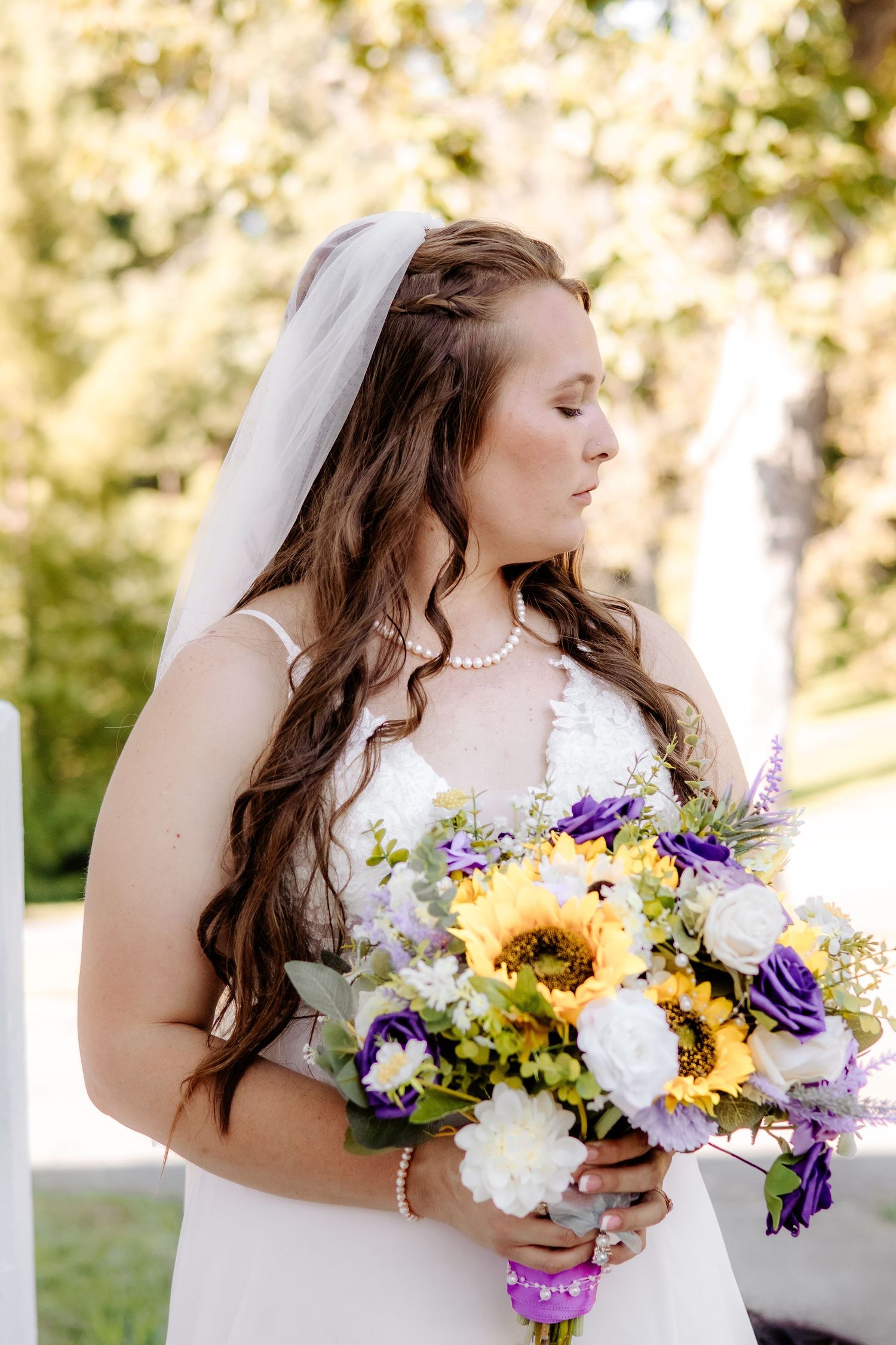 A bride in a white lace gown, veil and pearl necklace holds a bouquet of sunflowers, purple blooms and white flowers outdoors.