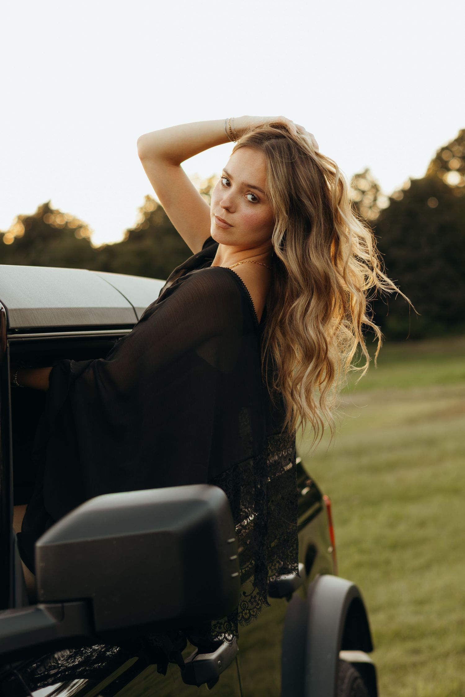 Woman with long wavy blonde hair leans out of a black Jeep window, one hand raised to her head, in a sunlit grassy field at golden hour.