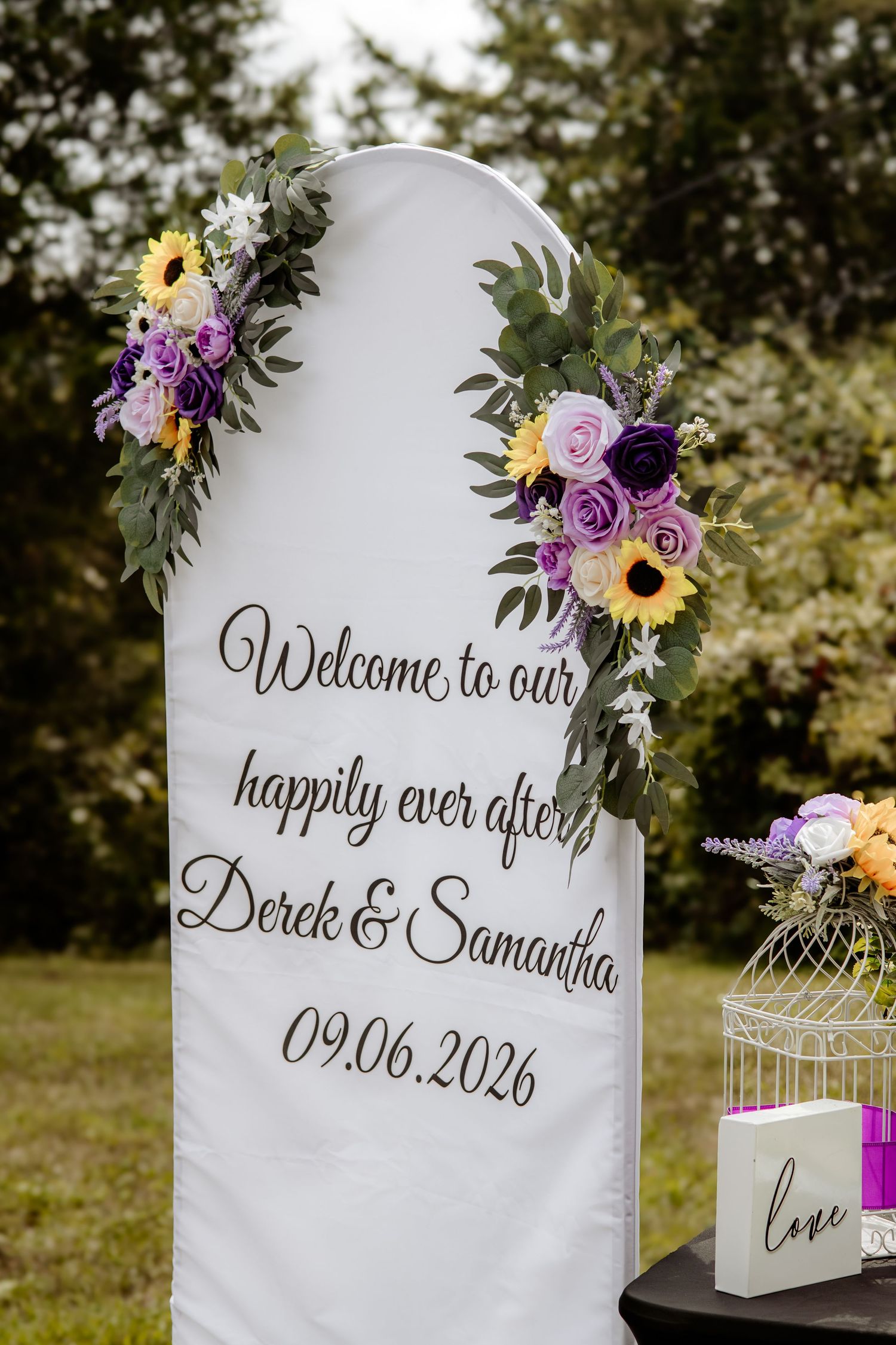 White wedding welcome sign with purple and yellow floral accents for Derek & Samantha, dated 09.06.2026, beside a birdcage and love sign.