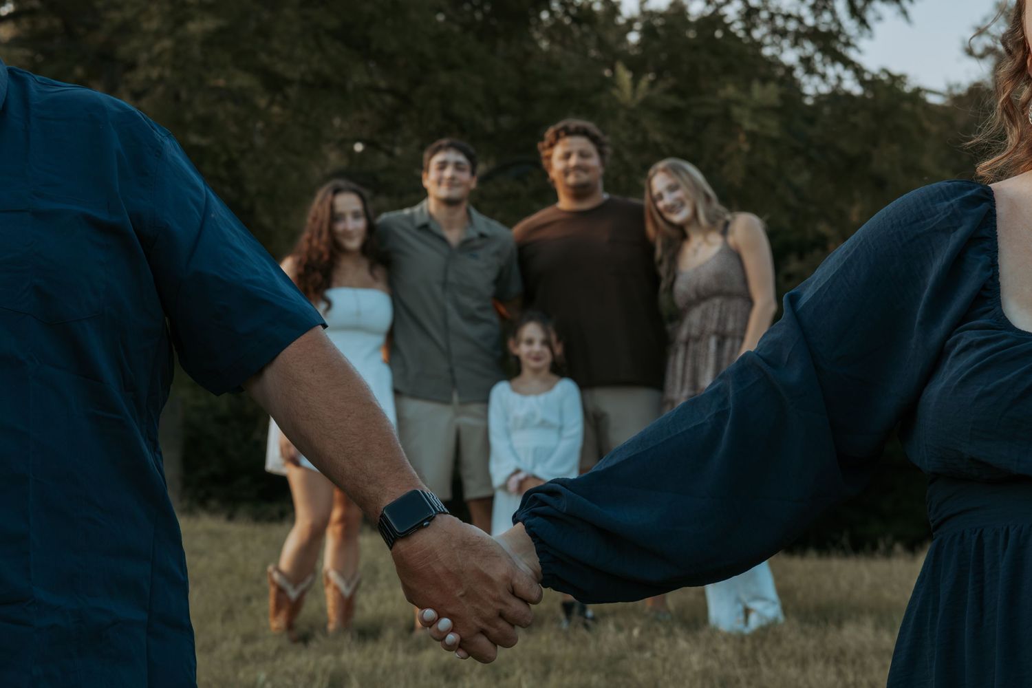 Family of six standing together in a grassy field at sunset while two people in the foreground hold hands.
