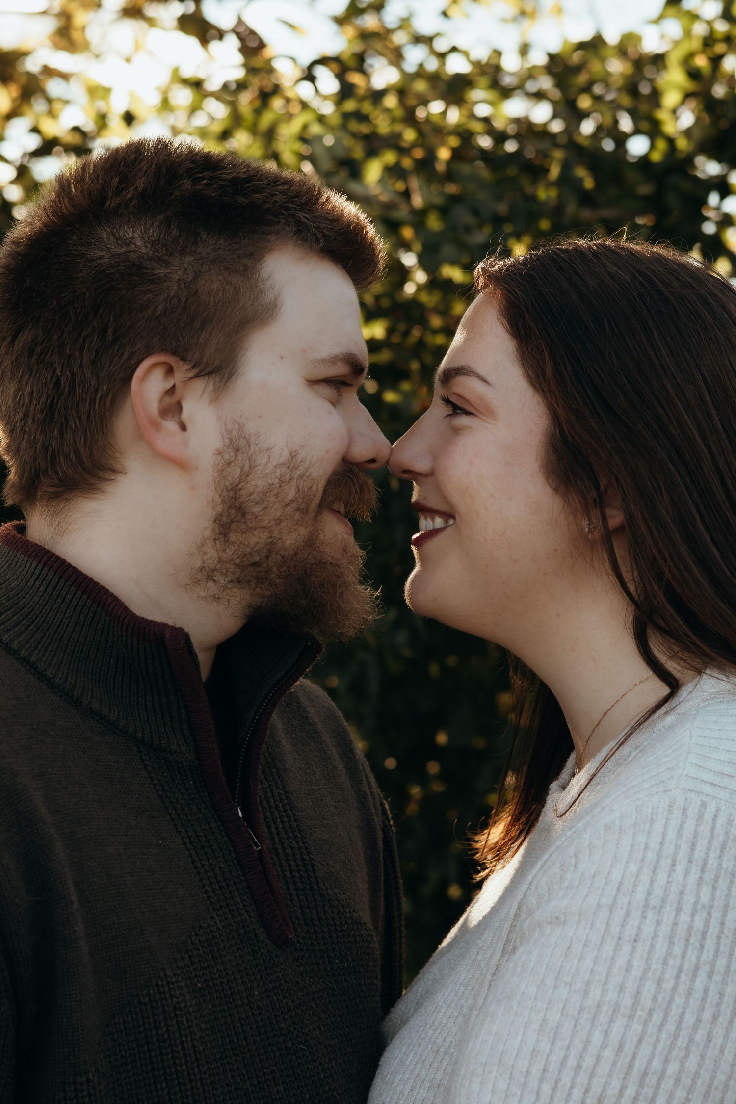 Smiling bearded man and woman with long dark hair touch noses in a romantic couple portrait against sunlit green foliage.