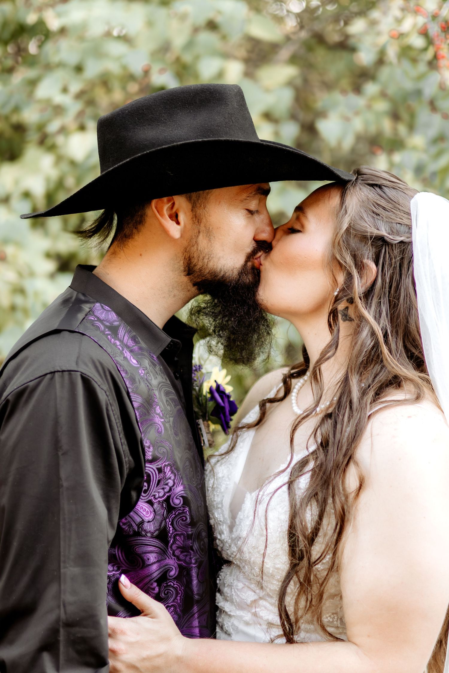 Bride in a lace gown and veil kisses a groom in a black cowboy hat and purple paisley vest during an outdoor wedding.