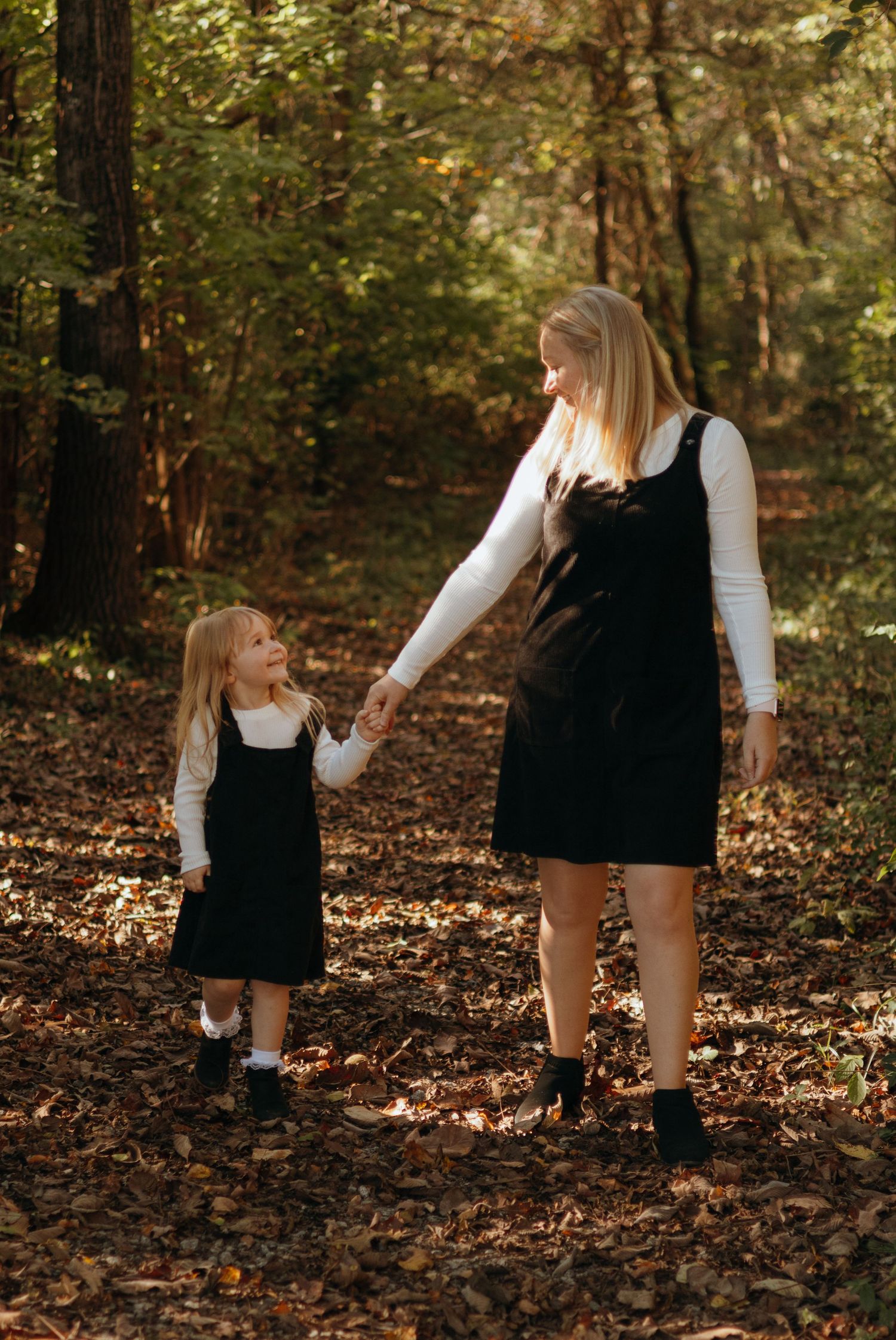 Woman and little girl in matching black pinafore dresses and white tops hold hands while walking on a leaf-covered forest path.