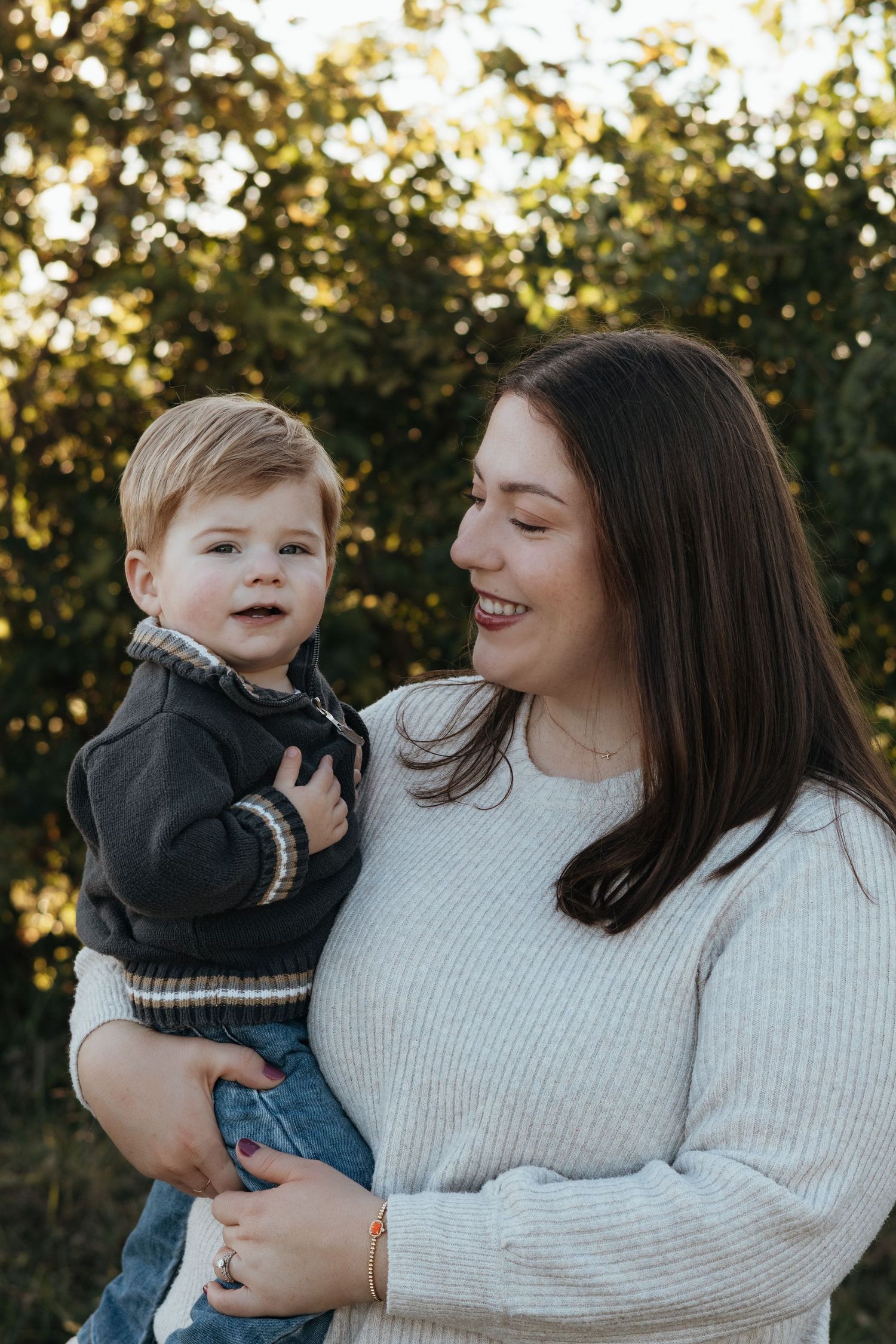 A smiling woman with long dark hair holds a toddler in a gray jacket outdoors, with warm sunlit green foliage glowing in the background.
