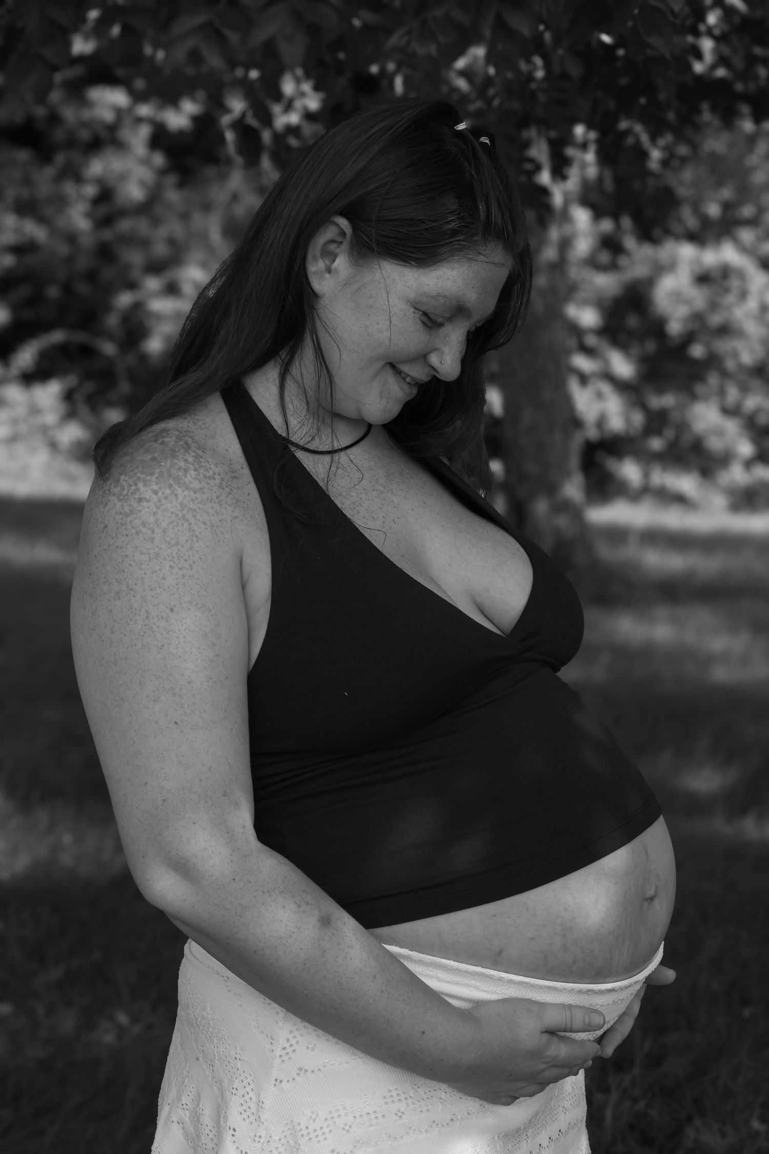 Black and white portrait of a smiling pregnant woman gently cradling her bare belly while standing outdoors beneath a tree.