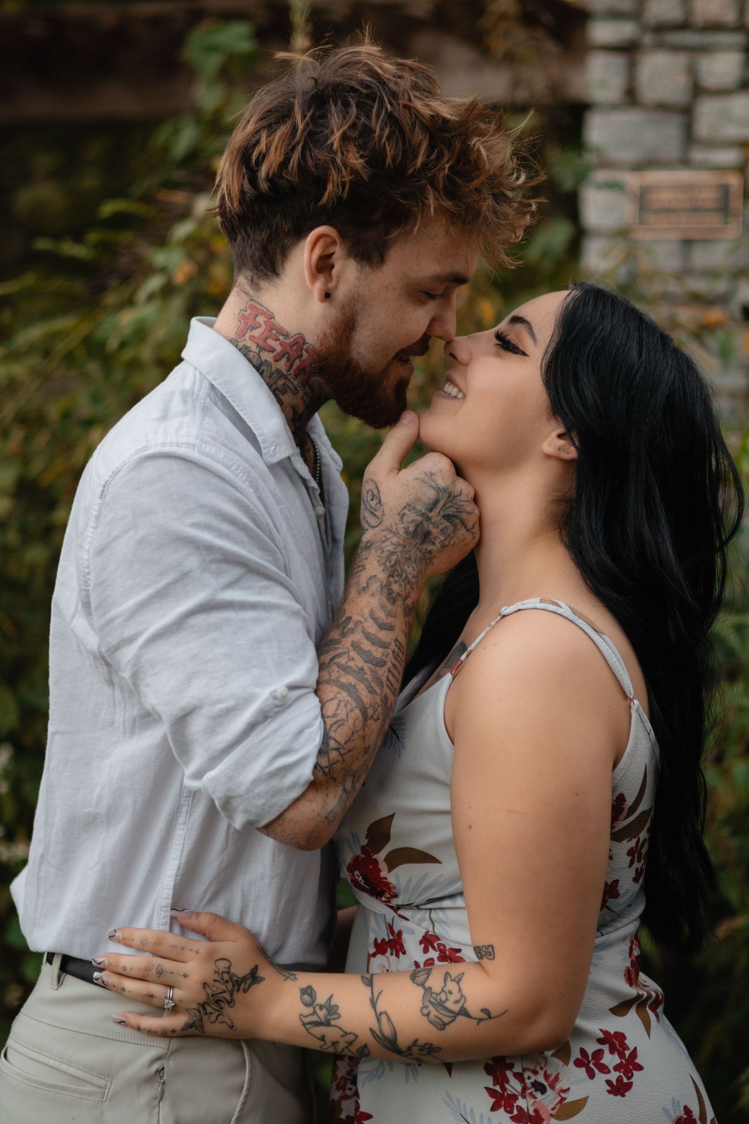 A tattooed couple shares an intimate moment outdoors, the man gently lifting the smiling woman's chin as she wears a floral dress.