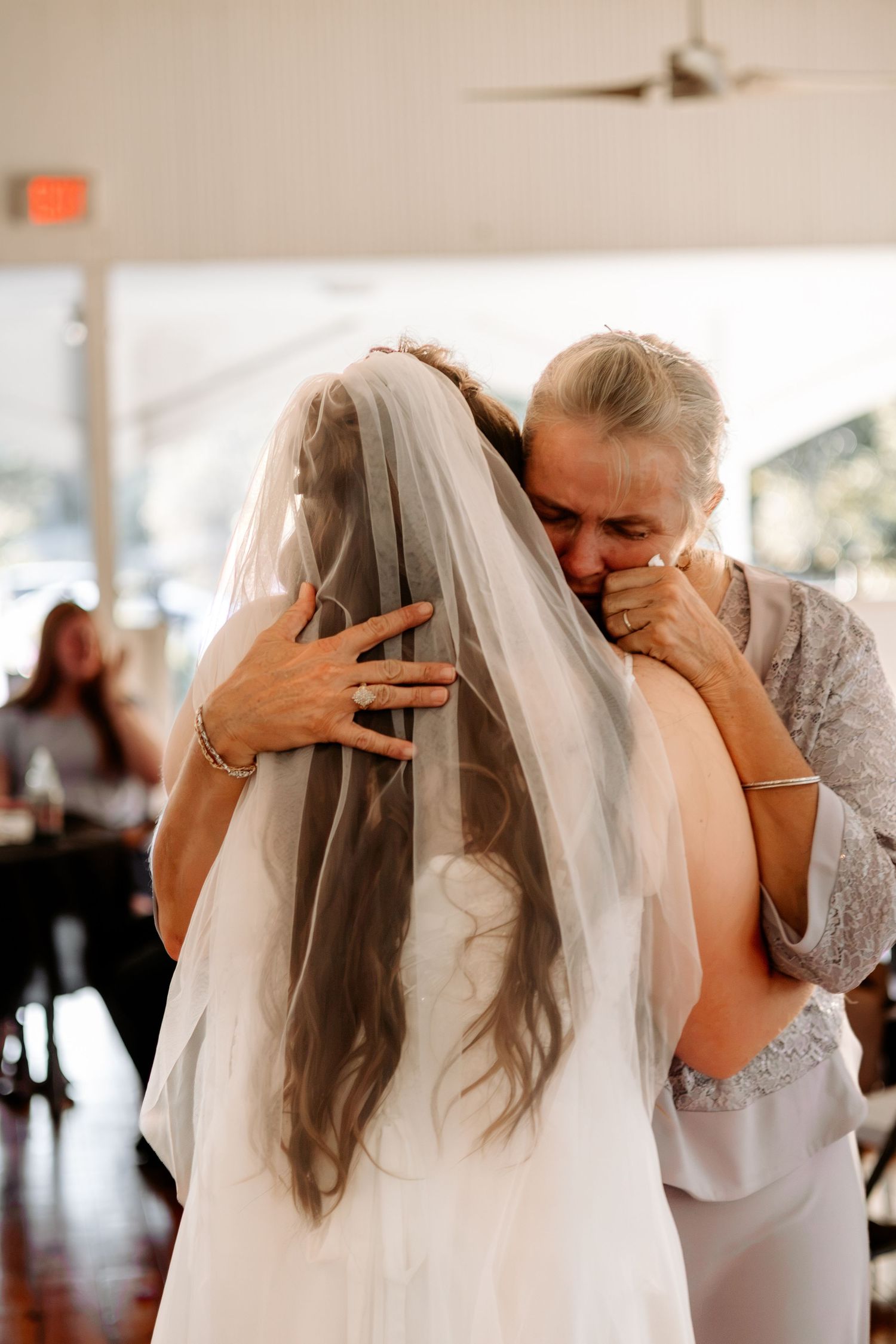 A bride in a long white veil shares an emotional embrace with an older woman during a wedding reception.