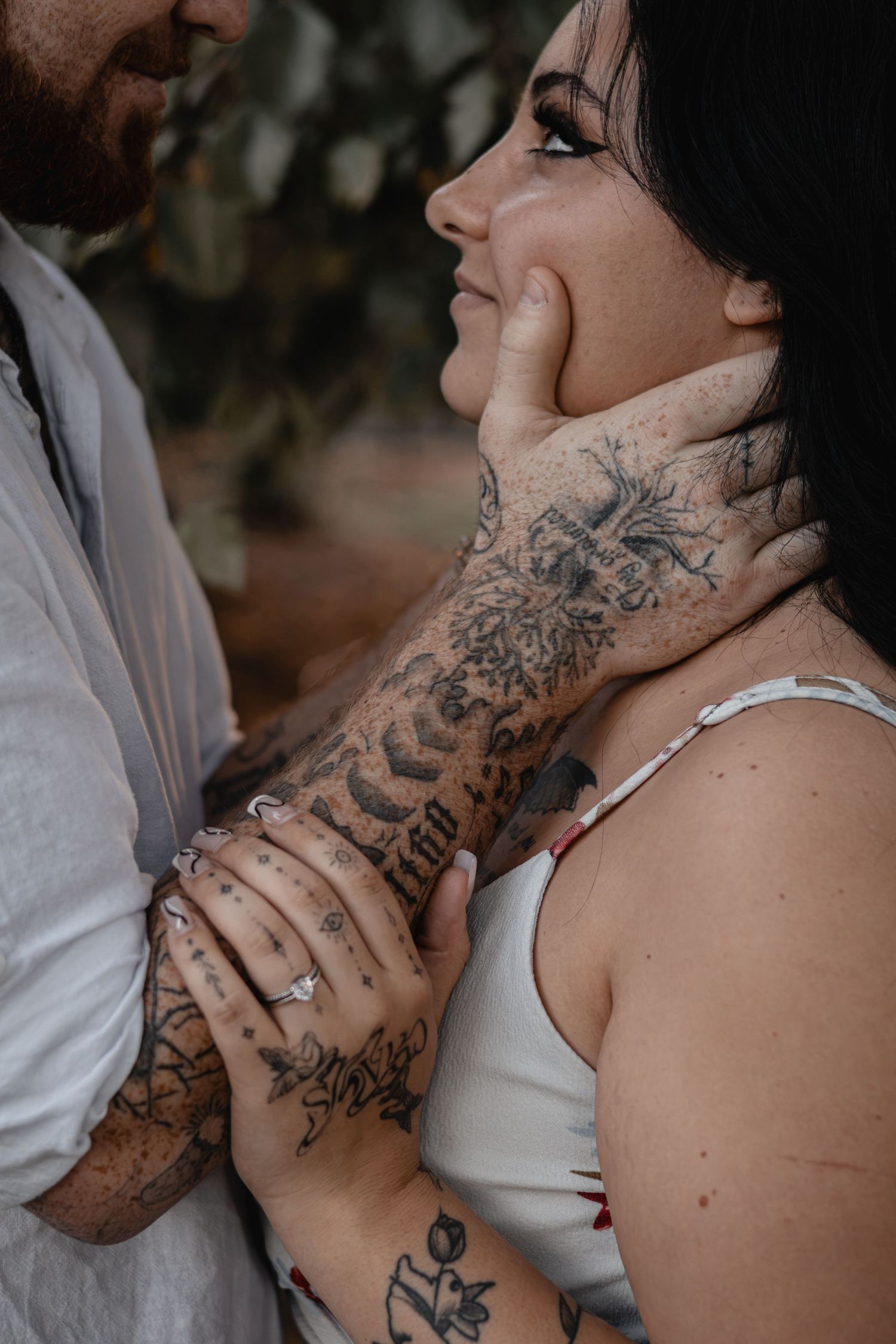 Close-up of a tattooed couple embracing, her face gently cradled by his inked hand, with a diamond ring visible in soft natural light.