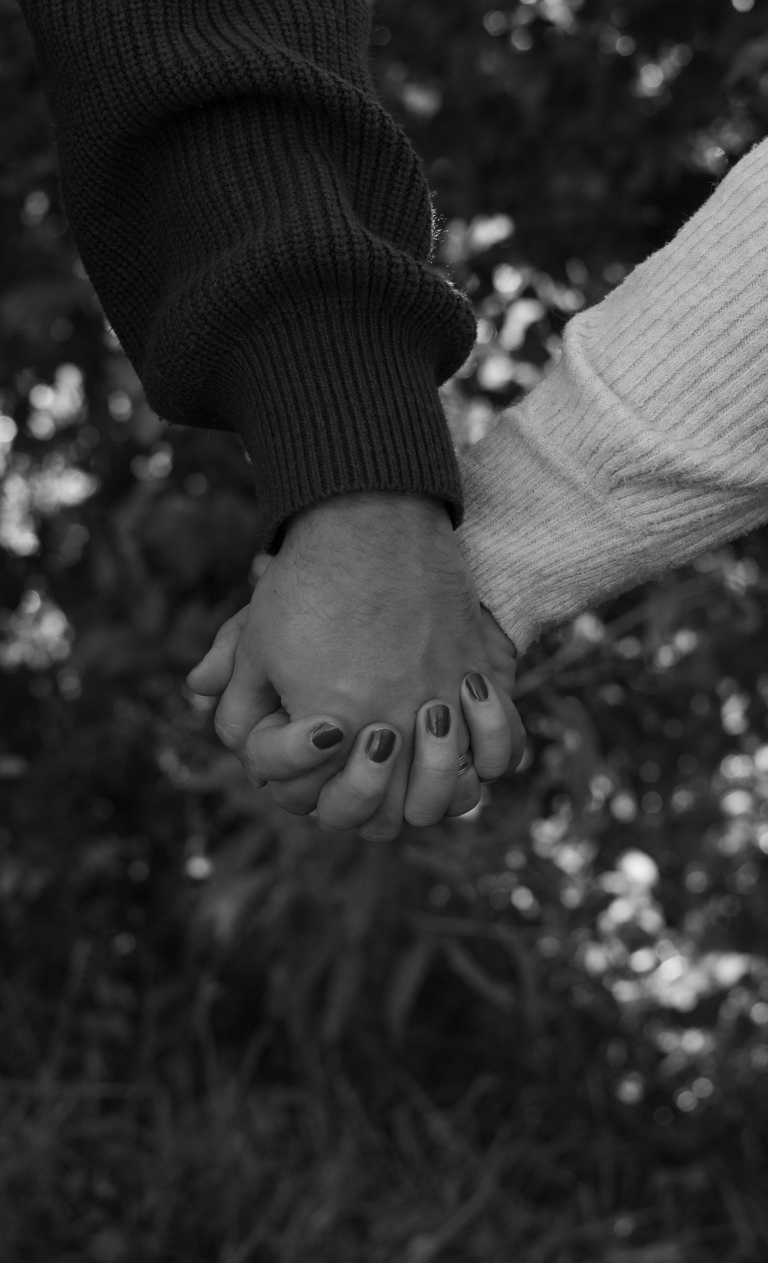 Black and white photo of a couple holding hands outdoors, wearing cozy sweaters with dark painted nails and a ring.