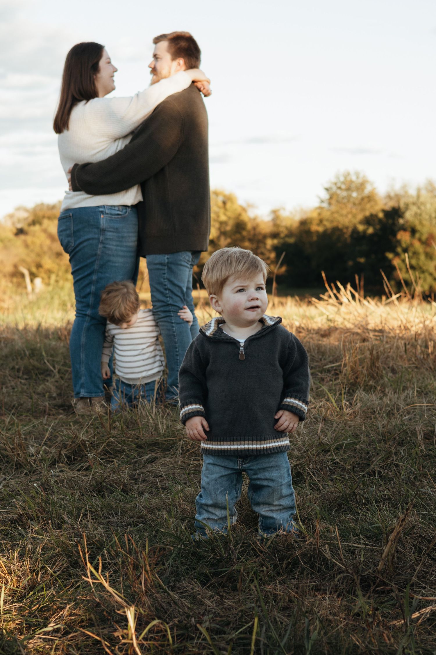 Toddler in a gray hooded sweater stands in a golden grassy field while his parents embrace and another child plays behind them at sunset.