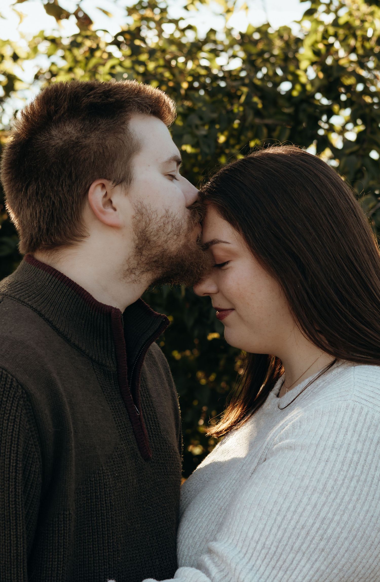 A bearded man in a green quarter-zip sweater tenderly kisses the forehead of a woman in a white sweater outdoors in golden sunlight.