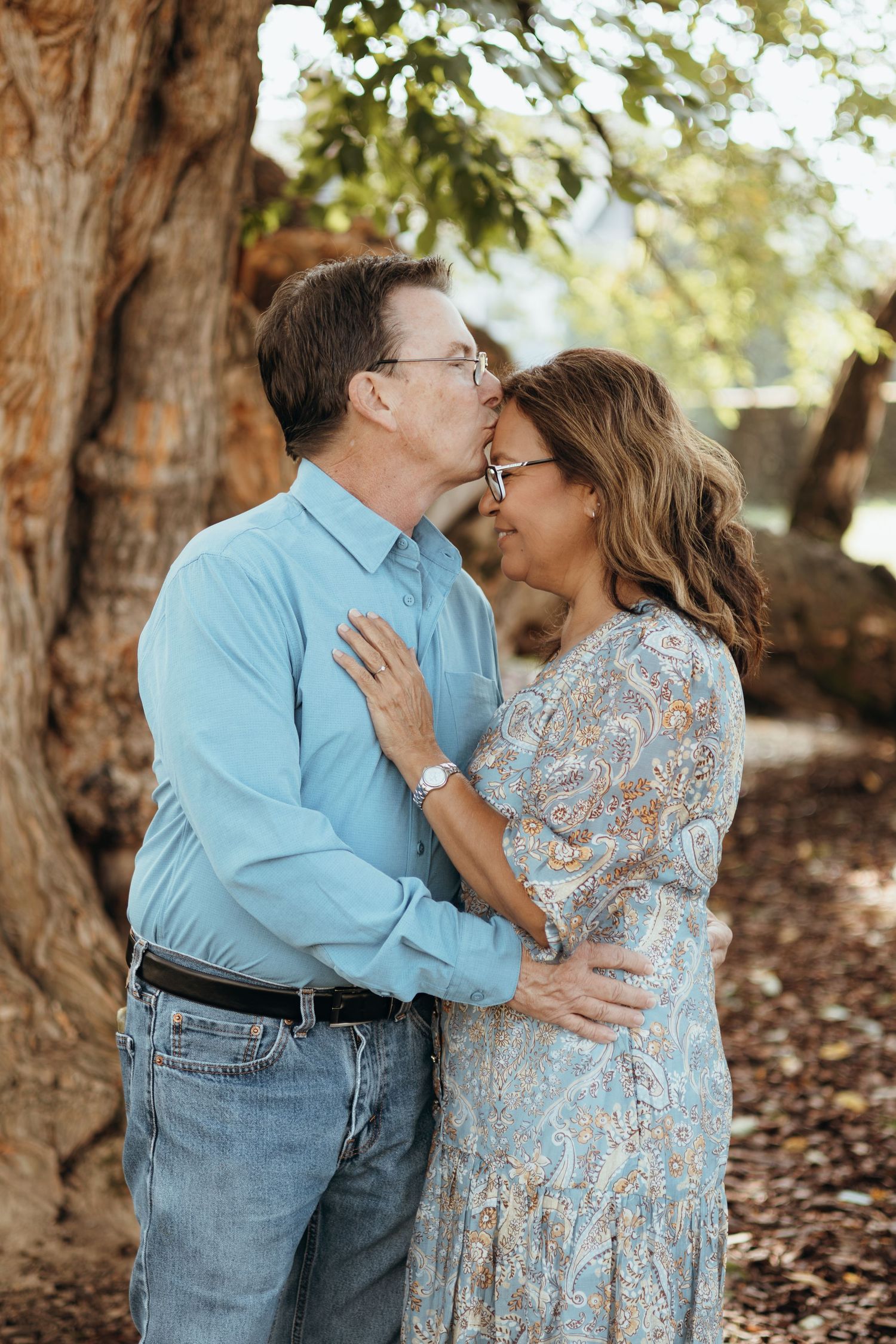 A man in a blue button-up shirt kisses the forehead of a smiling woman in a floral dress as they embrace beside a large tree.