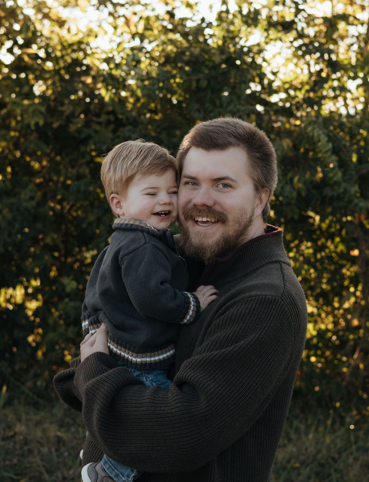 Smiling bearded man in a dark sweater holds a laughing toddler in a navy jacket outdoors with sunlit green trees in the background.